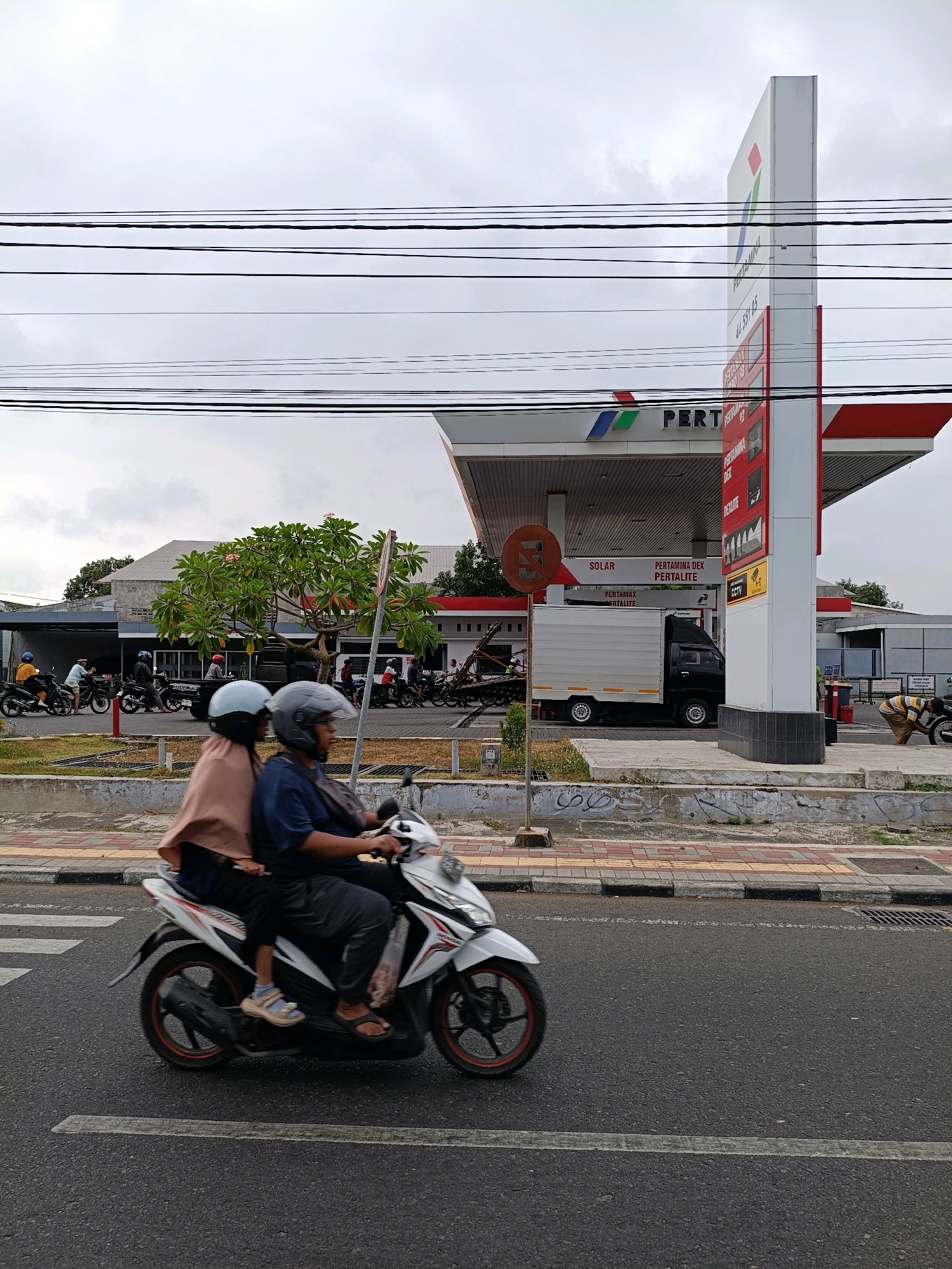 In Yogyakarta, Indonesia, a cloudy day unfolds at a Pertamina gas station. Two individuals, wearing helmets and dressed in casual attire, ride a white scooter. The person in front, wearing a dark blue shirt and black pants, is driving, while the person behind, wearing a light brown hijab and dark pants, is a passenger. They are positioned on the right side of the frame, moving from left to right across the asphalt road.
The gas station features a prominent white structure with red accents and the Pertamina logo. Red and white signs indicating fuel prices are visible on a tall, vertical pylon. Underneath the covered area of the station, a white truck with a black cab is parked. Several other motorbikes are visible in the background, suggesting a busy area. A green tree with pink flowers stands to the left of the truck. The sidewalk is paved with decorative tiles in red and yellow, bordered by grey concrete. Overhead, a complex network of electrical wires crisscrosses the sky, typical of urban Indonesian landscapes. The overall scene depicts a slice of daily life in a bustling urban environment.