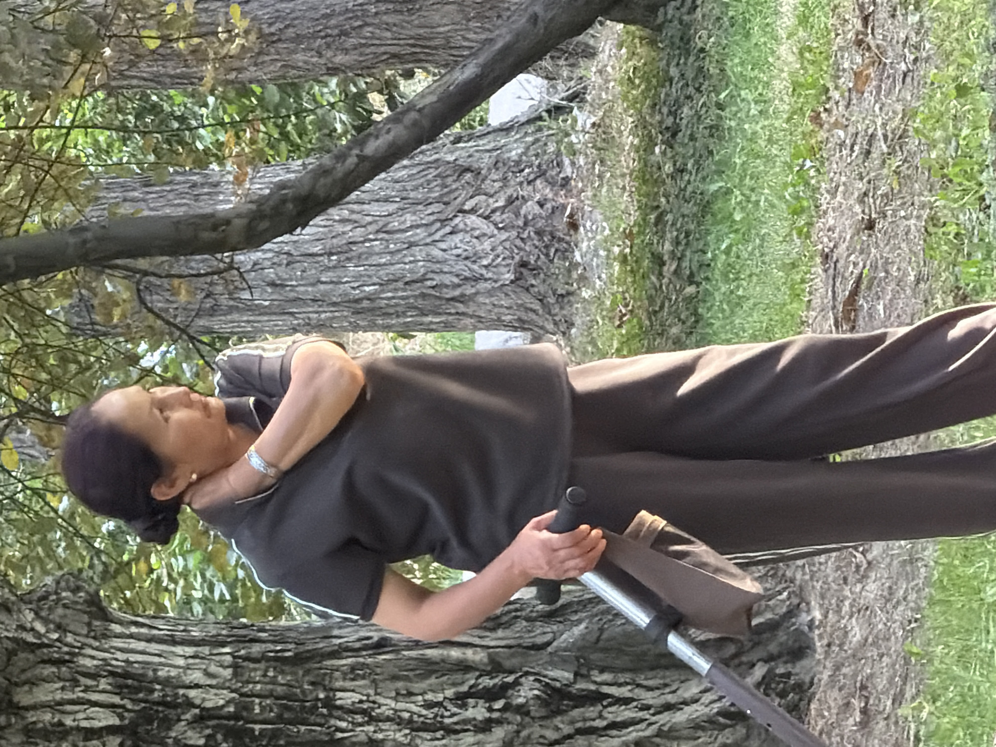A medium shot captures a woman of Asian descent from the knees up, standing in a park. She has dark hair pulled back and is wearing a dark grey short-sleeved polo shirt with white stripes on the collar and sleeves, paired with dark grey pants. Her right hand is touching her neck, and she wears a silver bracelet on her left wrist and a small earring in her left earlobe. The woman is holding the handle of a stroller with her right hand. The background consists of large trees with rough bark, some foliage, and green grass with scattered leaves and twigs on the ground. The lighting suggests it is daytime with dappled sunlight filtering through the trees. The woman is looking to her right, away from the camera.