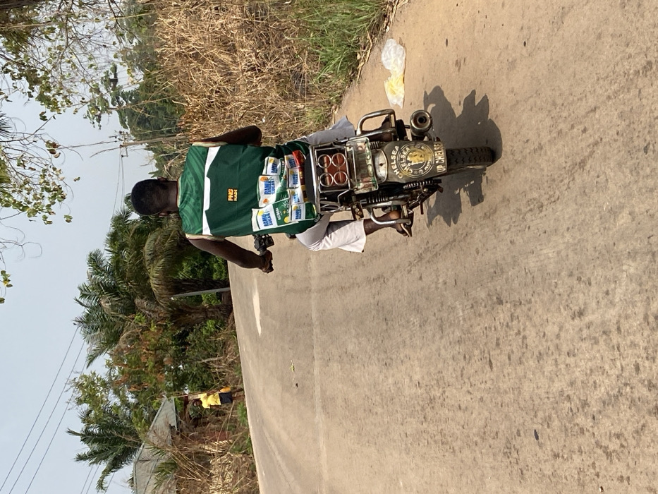 The image depicts a rural or semi-rural road scene in Igbarra, Nigeria, under bright daylight. The primary subject is an individual riding a dark-colored motorcycle away from the viewer, moving up a slight incline on a light brown, possibly unpaved or poorly paved road.
The motorcyclist is dark-skinned, wearing a green and white sleeveless top with "PNG MADE" visible on the back, white capri-length trousers, and dark sandals. The motorcycle features a rear rack laden with several white packages, prominently displaying "RAMU SUGAR" and possibly other "RAMU" branded products. The rear mudguard of the motorcycle is customized with a blue and gold emblem featuring "CHELSEA" and "UP CHELSEA" text. A single exhaust pipe is visible on the right side of the motorcycle's rear.
On the left side of the road, in the mid-distance, a second individual is visible. This person, appearing to have lighter skin, wears a yellow shirt and dark shorts, standing near a modest structure with a visible roofline and surrounded by dry, bushy vegetation. They are holding what appears to be a long pole or stick.
The immediate roadside environment on the right features dry, tall grass and shrubs. A crumpled white plastic bag rests on the shoulder of the road. Further back, numerous palm trees and other dense green foliage are visible under a clear, pale blue sky with some scattered high clouds. Utility lines are faintly visible across the upper portion of the frame.