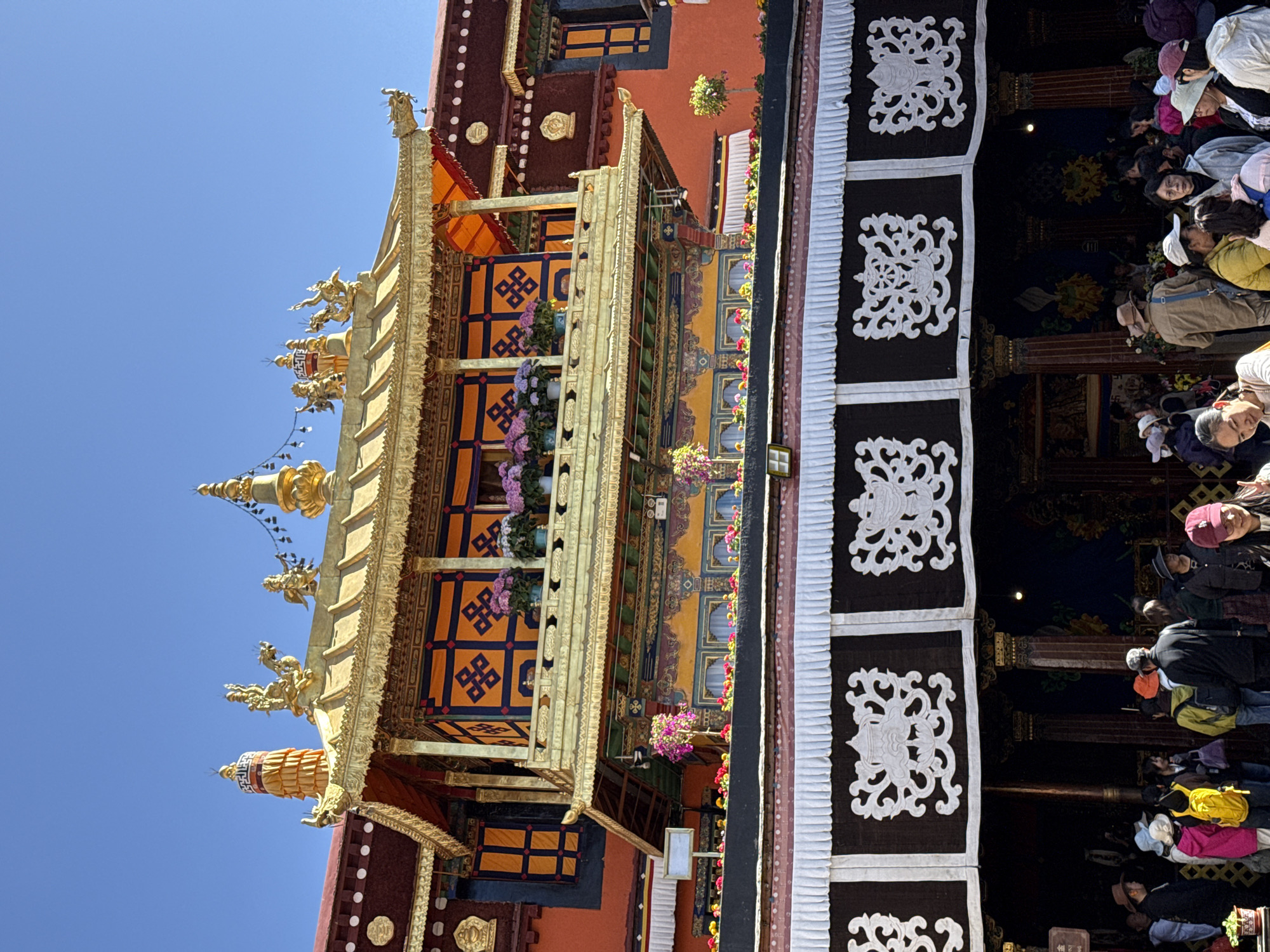 The Potala Palace, located in the city of Chengguan District, China, is depicted in an eye-level outdoor shot. A part of the palace's elaborate structure is in view, showcasing intricate golden decorations, colorful fabrics, and floral arrangements. A crowd of people is seen on the right side of the image, appearing to be tourists or visitors exploring the site. The sky above is clear and blue.