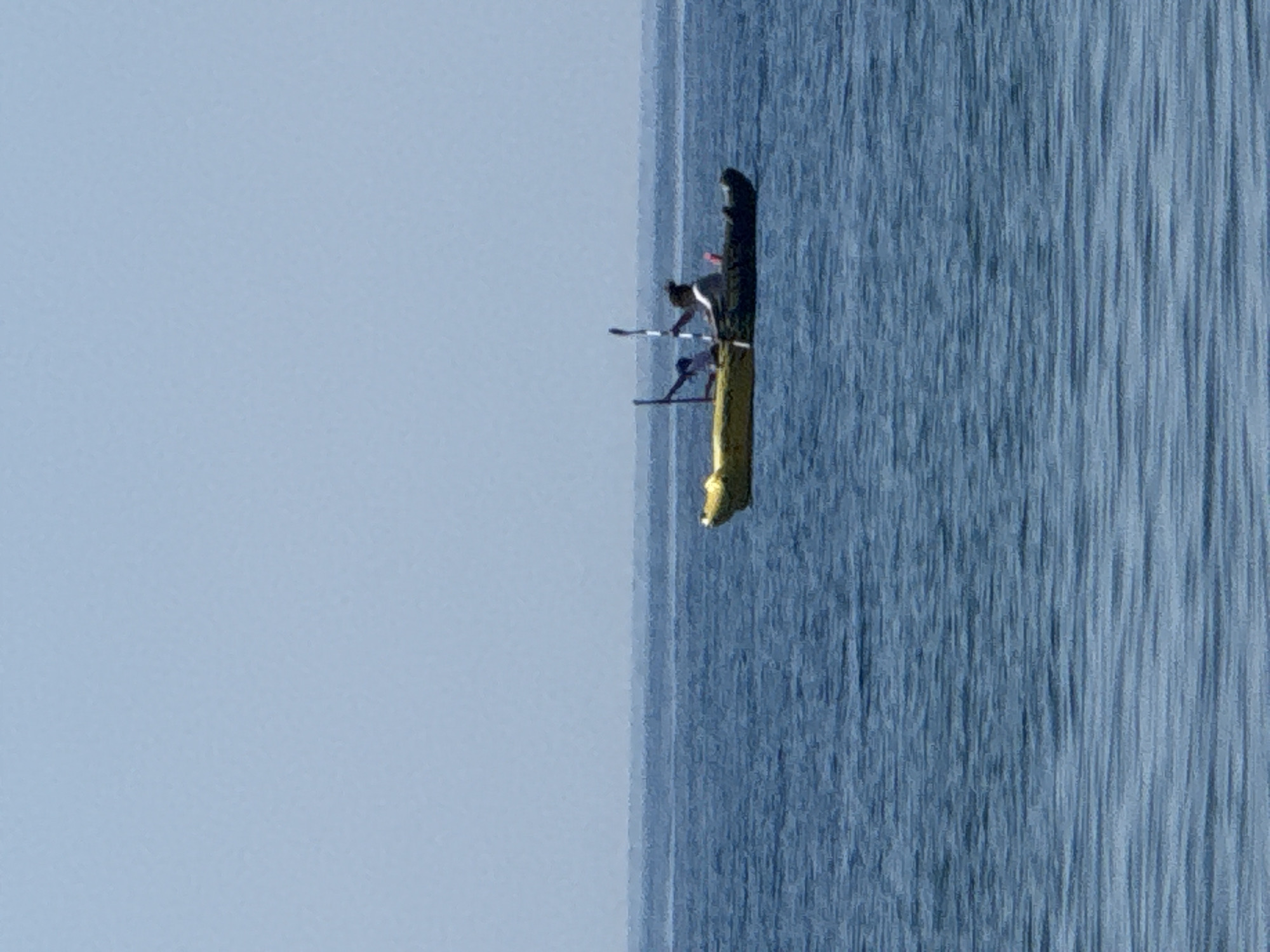 An aerial shot captures two individuals in a tandem kayak navigating the waters off the coast of Buljarica, Montenegro. The kayak, a two-person vessel, is predominantly yellow, with a darker section towards the rear. One person, positioned in the front of the kayak, is partially obscured, their presence indicated by a dark object, possibly an arm or paddle. The individual in the rear is more clearly visible, wearing a light-colored shirt and dark pants, and actively paddling with a two-bladed oar. They are looking forward, seemingly focused on steering. The kayak is floating on a calm, blue sea, with the horizon line clearly defined by a hazy, lighter blue sky. The water exhibits subtle ripples, suggesting a gentle breeze. The overall scene is tranquil, with the vast expanse of the sea and sky emphasizing the small scale of the kayak and its occupants.