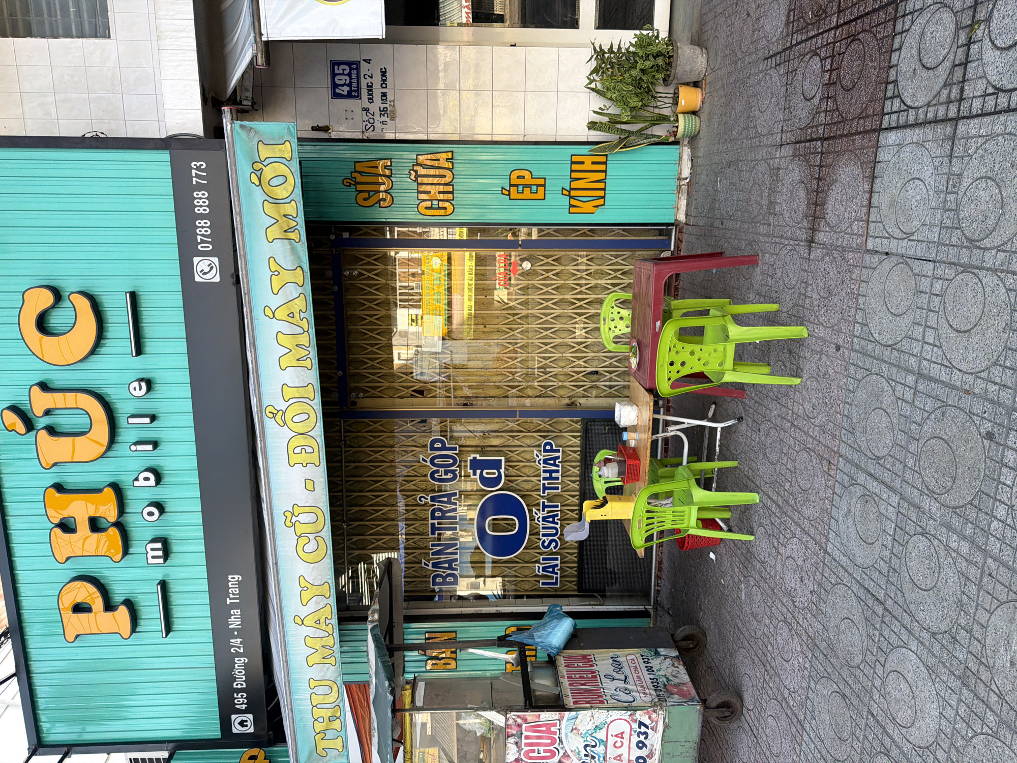 The image depicts a street scene in Bac Nha Trang Ward, Vietnam. A storefront with a prominent turquoise facade features the signage "PHÚC mobile" in large orange letters. Below this, a black awning displays an address "495 Đường 2/4 - Nha Trang" and a phone number "0788 888 773". A banner above the entrance reads "THU MÁY CŨ - ĐỔI MÁY MỚI" (Trade in old phones - exchange for new phones).
The storefront's glass doors are partially obscured by a yellow metal security gate. Inside, signs indicate services like "BÁN TRẢ GÓP" (Installment sales) and "LÃI SUẤT THẤP" (Low interest rates). To the right of the entrance, a narrow turquoise metal door has vertically aligned orange lettering spelling out "SỬA CHỮA ÉP KÍNH" (Repair, glass pressing).
In front of the store, a red plastic table is set with three bright green plastic chairs. A small plate with food is on the table. Two more green chairs are positioned around the table. A yellow portable tool or dispenser is also on the table. To the left of the entrance, a food cart with colorful signage is visible, including the text "BÚN RIÊU GIÁ CỐ LÚA" and phone numbers. Several potted plants, including snake plants and leafy green plants, are arranged to the right of the turquoise door. The ground is paved with square grey tiles, some with circular patterns. The overall scene suggests a busy commercial area.