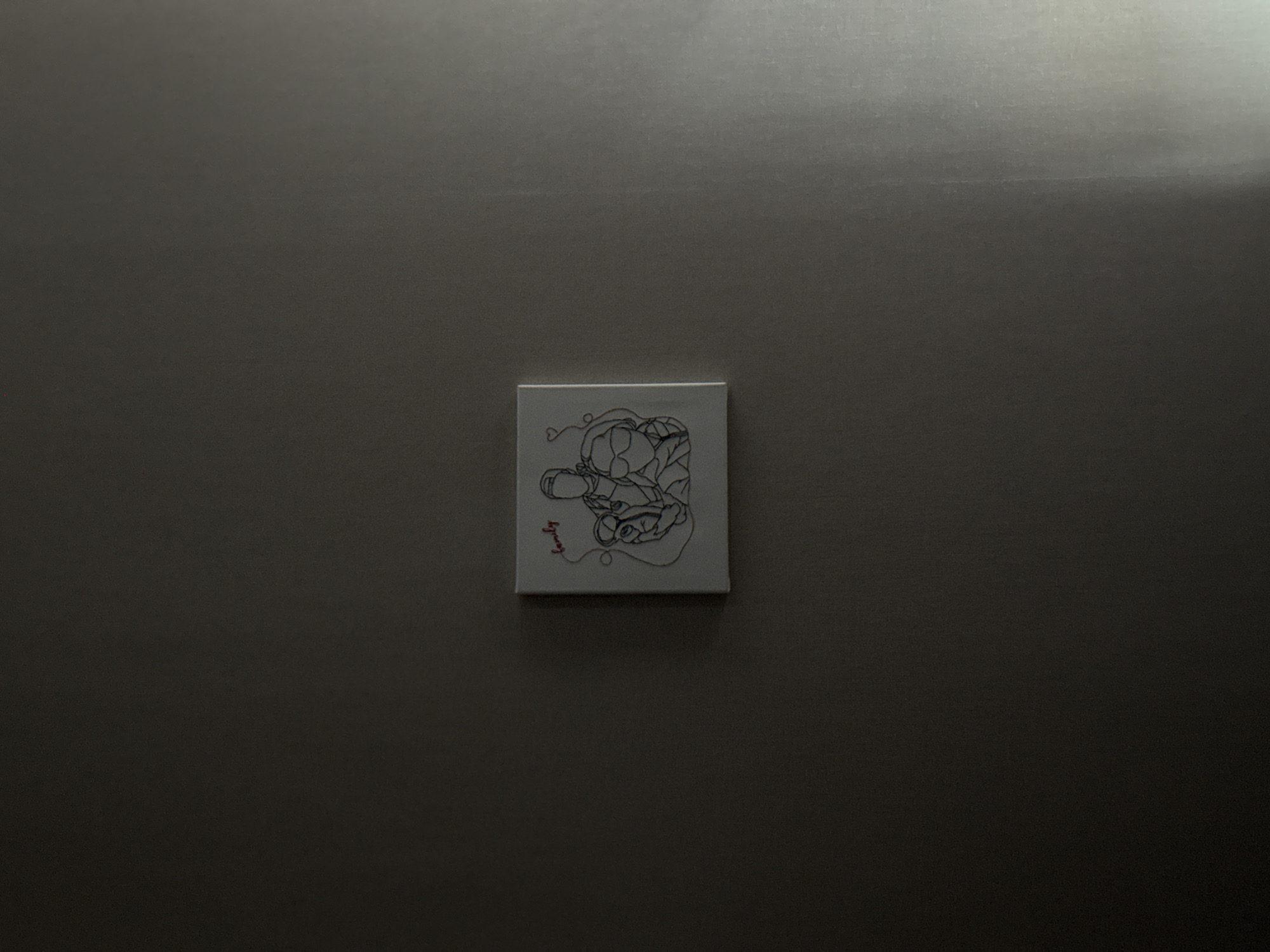 A small, square canvas with a white background hangs centered on a smooth, light-gray wall. The canvas features a black-line drawing, possibly an abstract depiction of figures or faces with connecting lines. To the lower left of the drawing, there is the word "family" written in red. A bright, vertical light source illuminates the wall from the upper right, creating a gradient effect and casting a slight shadow beneath the canvas. The location of the image is not discernible and cannot be identified as Izhevsk, Russia, or any other specific location.