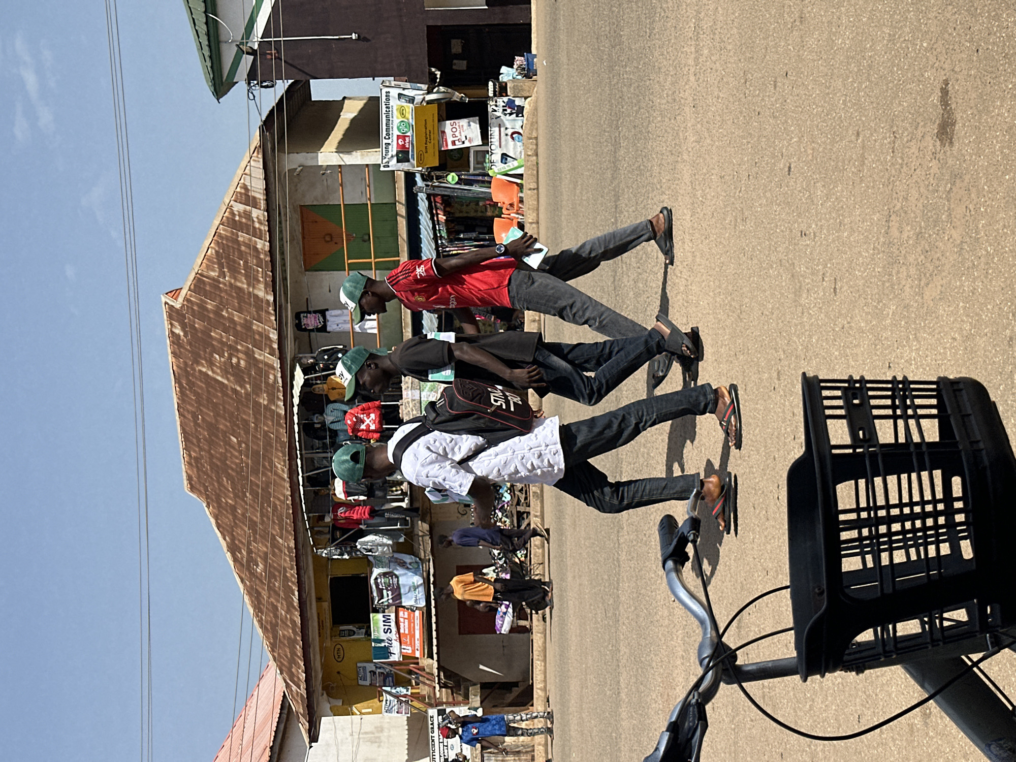 This media file shows a street scene in Bukuru, Nigeria. The image features several people walking along a paved road in front of shops. The shops appear to be selling clothing and possibly mobile phone services, as indicated by the visible text which includes "Da Young Communications," "SIM Registration Center," and brand names like "MTN" and "glo."
The subjects in the foreground are three young men, all wearing casual attire and some with baseball caps. They are walking in the same direction, with two of them appearing to be engaged in conversation. One man in a red shirt is holding a small item, possibly a pamphlet or phone. Another man is wearing a white patterned shirt and a backpack. In the background, more people are visible, some browsing in the shops and others walking on the street.
The scene suggests a typical daytime setting with bright sunlight, casting shadows on the ground. The weather appears to be clear and sunny. The architecture of the buildings is simple, with corrugated iron roofs visible on some structures, characteristic of the region. The overall atmosphere seems to be one of everyday activity, with people going about their business. The presence of various mobile network advertisements hints at a busy commercial area focused on telecommunications and related services.