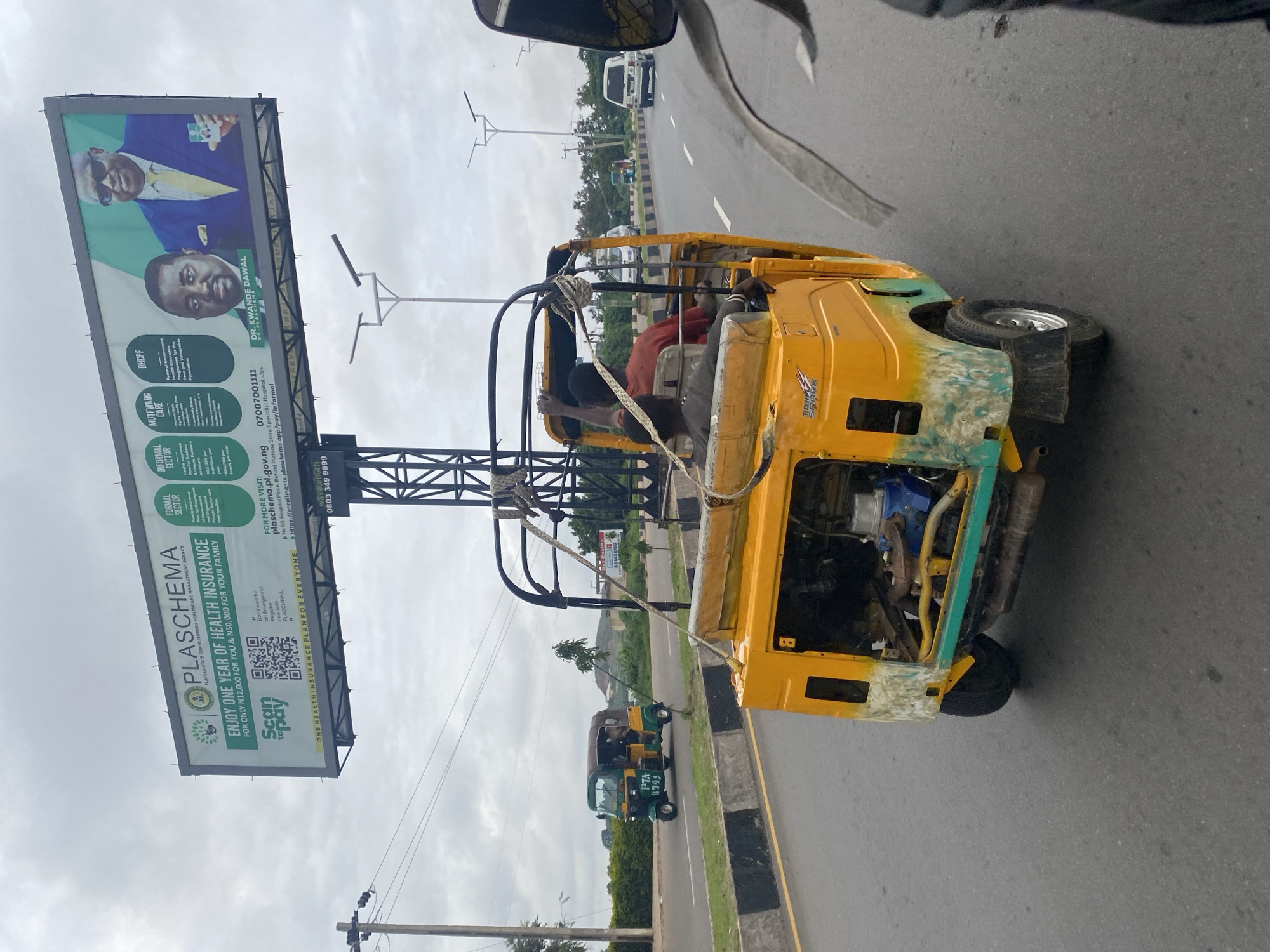 An outdoor, eye-level, medium shot shows a yellow, three-wheeled auto-rickshaw driving on a road in Jos, Nigeria. The auto-rickshaw is in the foreground and is partially obscuring the view of the road ahead. Two men are visible in the auto-rickshaw. One man, wearing a red shirt, is in the driver's seat. The other man, wearing a grey shirt, is seated behind the driver. The auto-rickshaw appears to be old and is showing signs of wear and tear. The engine is exposed, and the body of the vehicle is partially rusted and peeling. The auto-rickshaw has a yellow body with a green trim.
In the background, a large billboard for PLASCHEMA is visible. The billboard advertises health insurance plans and features images of two men, identified as Dr. Kwande Dawal and Dr. Plaschema. The billboard also includes a QR code and contact information for PLASCHEMA.
Other vehicles, including a green and yellow auto-rickshaw, a white van, and a silver car, are visible on the road in the distance. The sky is overcast, and the overall lighting suggests it is daytime. The environment appears to be a suburban or urban setting, with trees and vegetation lining the roadside. The road is paved and has lane markings. The overall impression is a glimpse of everyday life and transportation in Jos, Nigeria, with a prominent advertisement for health insurance in the background.