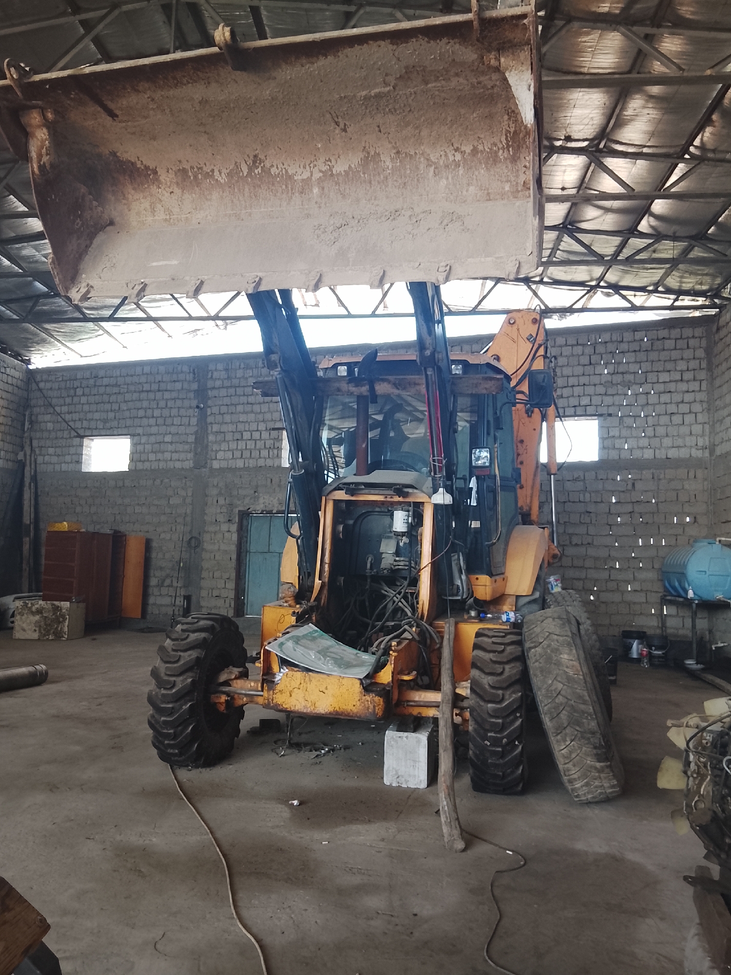 In a garage located in Sabir Rakhimov, Uzbekistan, a yellow backhoe loader is parked indoors. The machine has a large, dirty bucket raised high above its cab. The garage has a corrugated metal roof supported by a metal frame, and the walls are constructed of light-colored bricks. Two small windows are visible in the brick wall. In the background, there are cabinets, a blue door, and a blue water tank. Various objects, including tires and machine parts, are scattered around the garage floor. A white rope lies on the concrete floor, extending from the front of the backhoe. The scene appears to be a workshop or storage area for construction equipment.