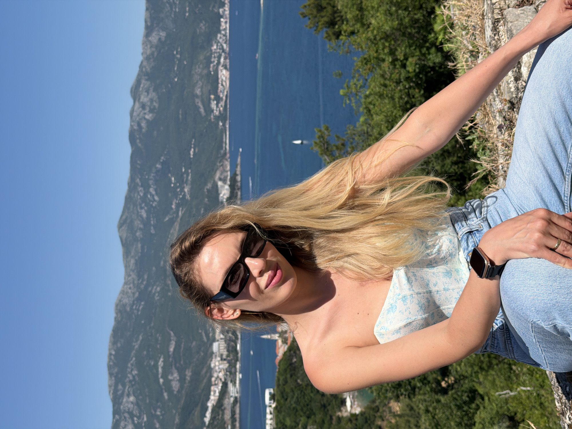 A woman with long blonde hair, wearing black rectangular sunglasses with "VOGUE" branding on the left temple, is depicted in a daytime outdoor setting. She is wearing a light blue and white floral strapless corset-style top and light blue denim jeans. Her left wrist displays a smartwatch with a rose gold-toned frame and a dark strap, and she wears a plain gold-colored ring on her left ring finger. She also has a small stud earring and a longer chain earring in her left ear. She is seated, with her left arm bent and resting on her leg, and her right arm extended to her side. Her lips are a light pink color, and she is looking towards the camera with a slight smile.
The background reveals a panoramic view of Budva, Montenegro. The deep blue Adriatic Sea occupies the midground, with a single white sailboat visible in the water, along with other smaller boats. The coastline features numerous buildings, including the red-tiled roofs and a prominent church spire indicative of Budva's Old Town, and modern structures stretching along the shore. Lush green mountains rise in the distance under a clear blue sky. In the immediate foreground and surrounding the woman are green foliage, dry grass, and rocks, suggesting an elevated viewpoint overlooking the bay.