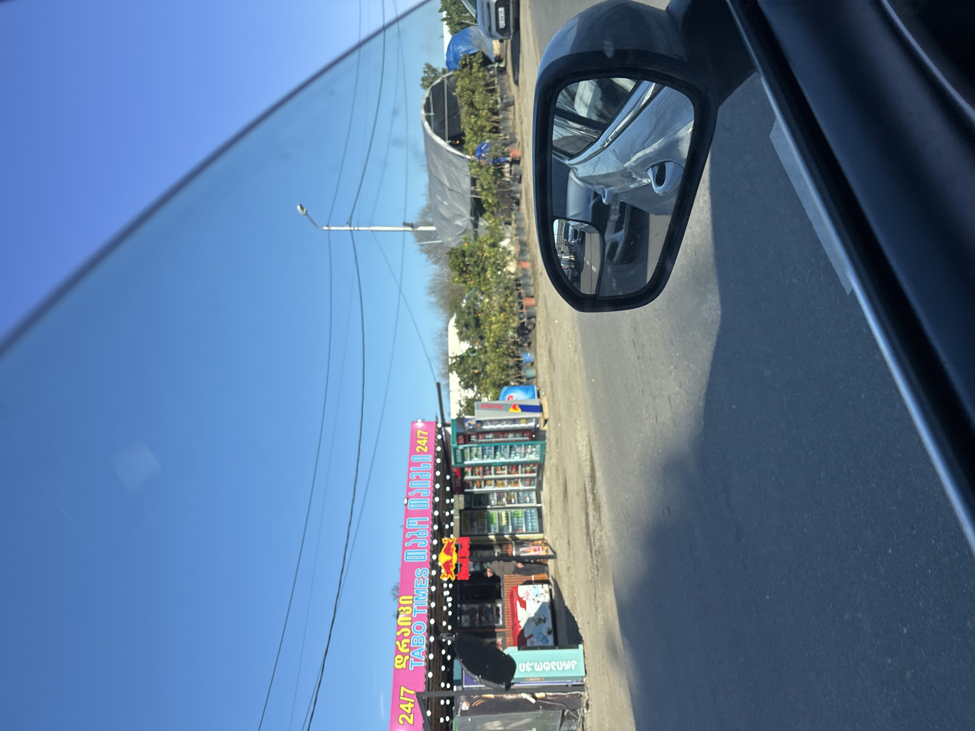 The image shows a view from inside a car, looking out. The scene captures a bright, clear day with a cloudless blue sky visible through the car window. Overhead power lines stretch across the sky. Alongside the road is a small retail building or kiosk identified by the pink sign displaying the text "TABO TIMES 24/7" in both English and Georgian script. The kiosk appears to be a small convenience store. In the car's side mirror, there is a reflection of another vehicle and a glimpse of a garden center with greenhouses and various plants. The car is driving on an asphalt road, typical for a city setting. Based on the signage in Georgian script, the scene is likely located in the city of Tbilisi, Georgia.