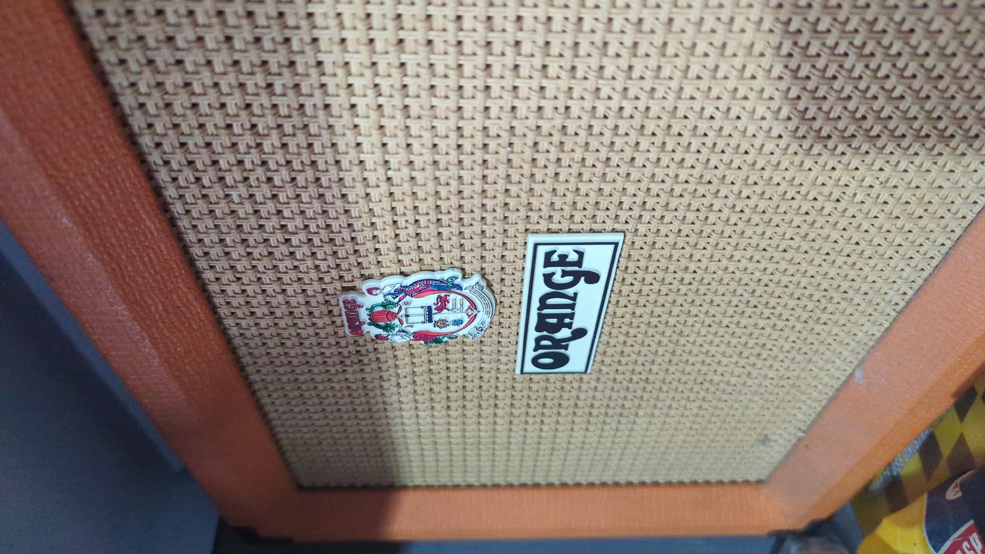 A close-up, eye-level shot focuses on the speaker grille of an Orange brand amplifier. The grille is a woven, textured material in a light tan color, with a tightly woven pattern. The amplifier's cabinet is trimmed in a textured orange material, visible along the left and top edges of the frame. Two distinct emblems are affixed to the grille. The upper emblem is a circular crest with a complex design featuring figures and symbols. Below it, a rectangular emblem displays the word "ORANGE" in bold, white, stylized lettering on a black background. The lighting in the scene is soft, casting subtle shadows that highlight the texture of the grille and cabinet. The image does not depict any specific location within Penjor, Indonesia, as it is a close-up product shot of an electronic device.