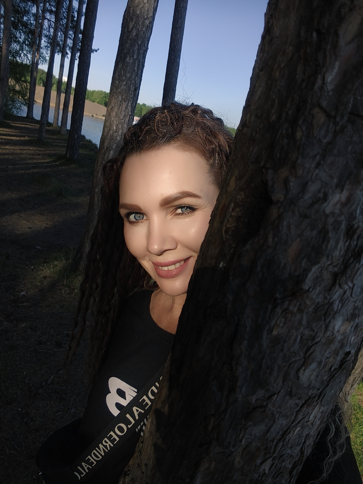 The image shows a woman standing behind a tree in a forested area, with a body of water visible in the background. The woman has fair skin, blue eyes, and curly brown hair. She is wearing a black t-shirt with a white logo. The trees are tall and slender, with dark bark. The sky is light blue. The location is in the city of Unknown, Unknown.