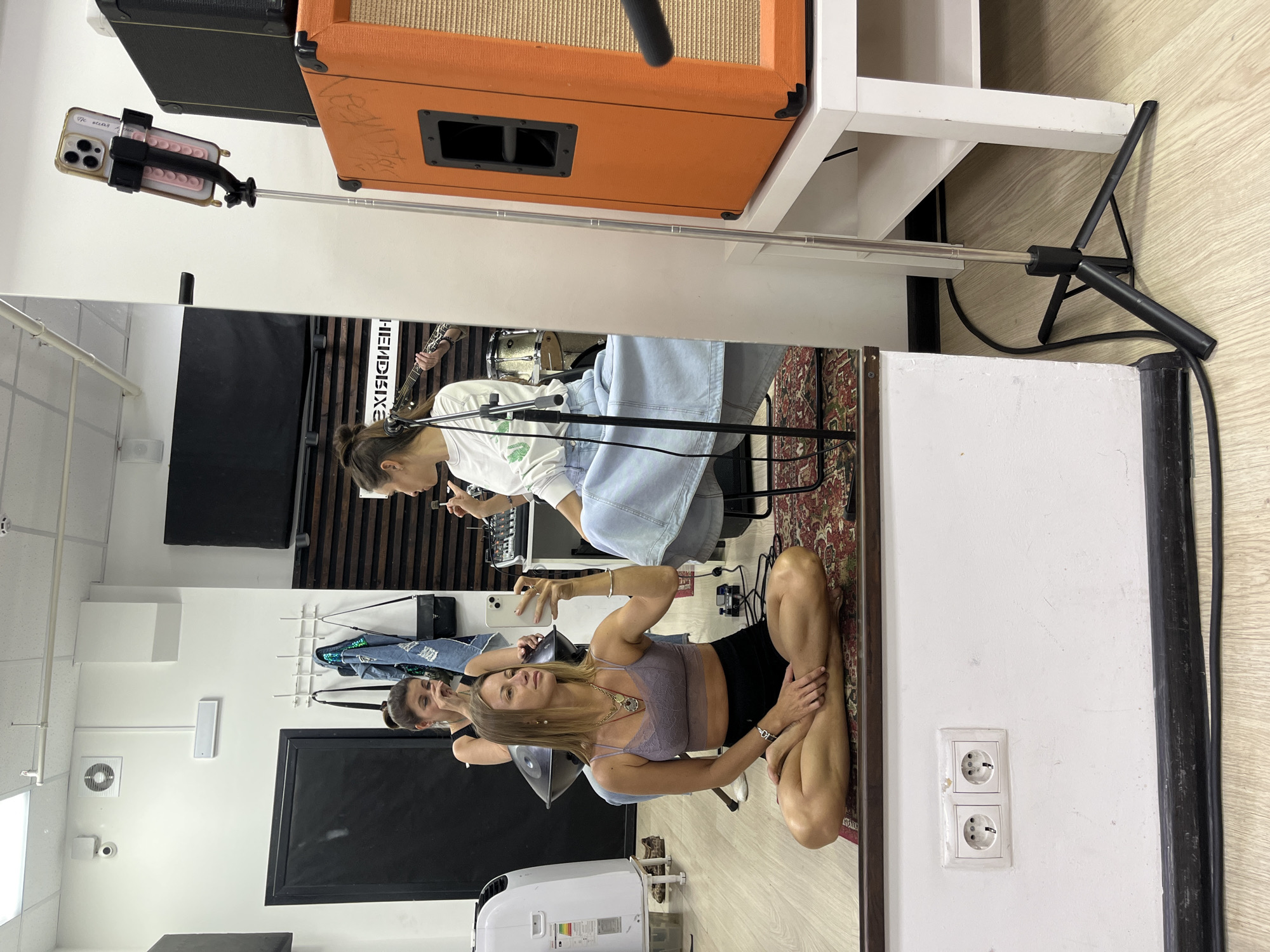 The image depicts an indoor scene with three women, likely in a music studio or rehearsal space. A woman in the foreground is sitting cross-legged on the floor in a yoga pose, wearing a grey bralette and black shorts, and holding a smartphone. She is looking into a mirror, as is another woman sitting behind her. This second woman is wearing a black top and appears to be touching her face. A third woman is visible in the background, also in the reflection of the mirror, sitting and playing a guitar. She is wearing a white long-sleeved shirt and a denim skirt. The environment includes a large mirror, sound equipment including an orange amplifier, and a drum set. The floor is wooden, with a patterned rug under the woman in the foreground. Electrical outlets are visible on the wall. The scene is likely in Moscow, Russia.