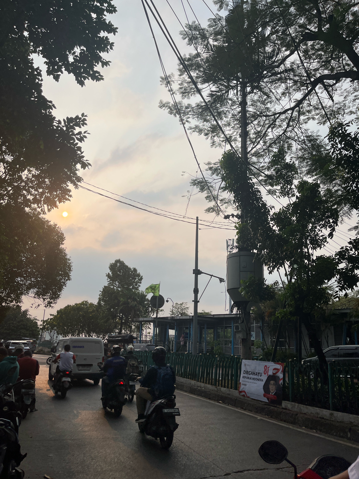 The image depicts a street scene in East Jakarta, Indonesia, during what appears to be either sunrise or sunset, given the low angle of the sun casting long shadows and the warm, diffused light. The foreground is dominated by a road with several motorcycles and a white van in transit. A few people are visible riding motorcycles, some with backpacks. On the right side of the road, a green fence runs along a sidewalk, behind which is a building with blue accents and a banner advertising "Dirgahayu Republik Indonesia" (Long Live the Republic of Indonesia) with the number 81 prominently displayed. Several trees with dense foliage are scattered throughout the scene, some silhouetted against the sky. The sky itself is a mix of pale blue and soft orange clouds, with the sun visible as a bright orb through a gap in the trees on the left. Utility poles with numerous wires are also present, stretching across the frame.