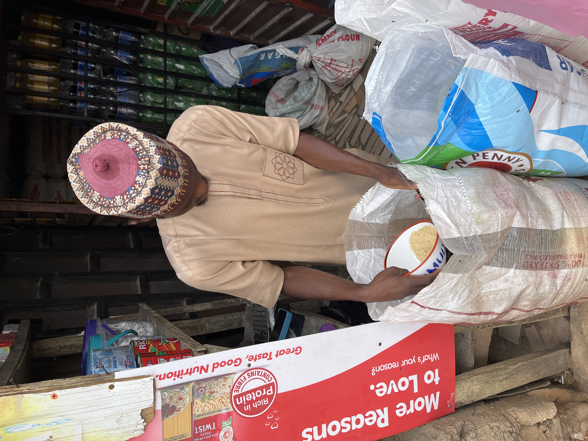 A man wearing a beige tunic and a colorful woven cap is seen in what appears to be a marketplace or store in Jos, Nigeria. He is in the process of scooping rice from a large white sack into a small white bowl. The sack is partially open, revealing a white and blue patterned design. Behind him, more sacks of flour and sugar are visible, along with shelves stocked with bottled drinks. In the foreground, a red and white sign displays text in English, including "More Reasons to Love," "What's your reason?", "Great Taste, Good Nutrition," and "Rich in Protein." The scene suggests a commercial environment, likely a foodstuff vendor's stall. The lighting is natural, indicating it is daytime.