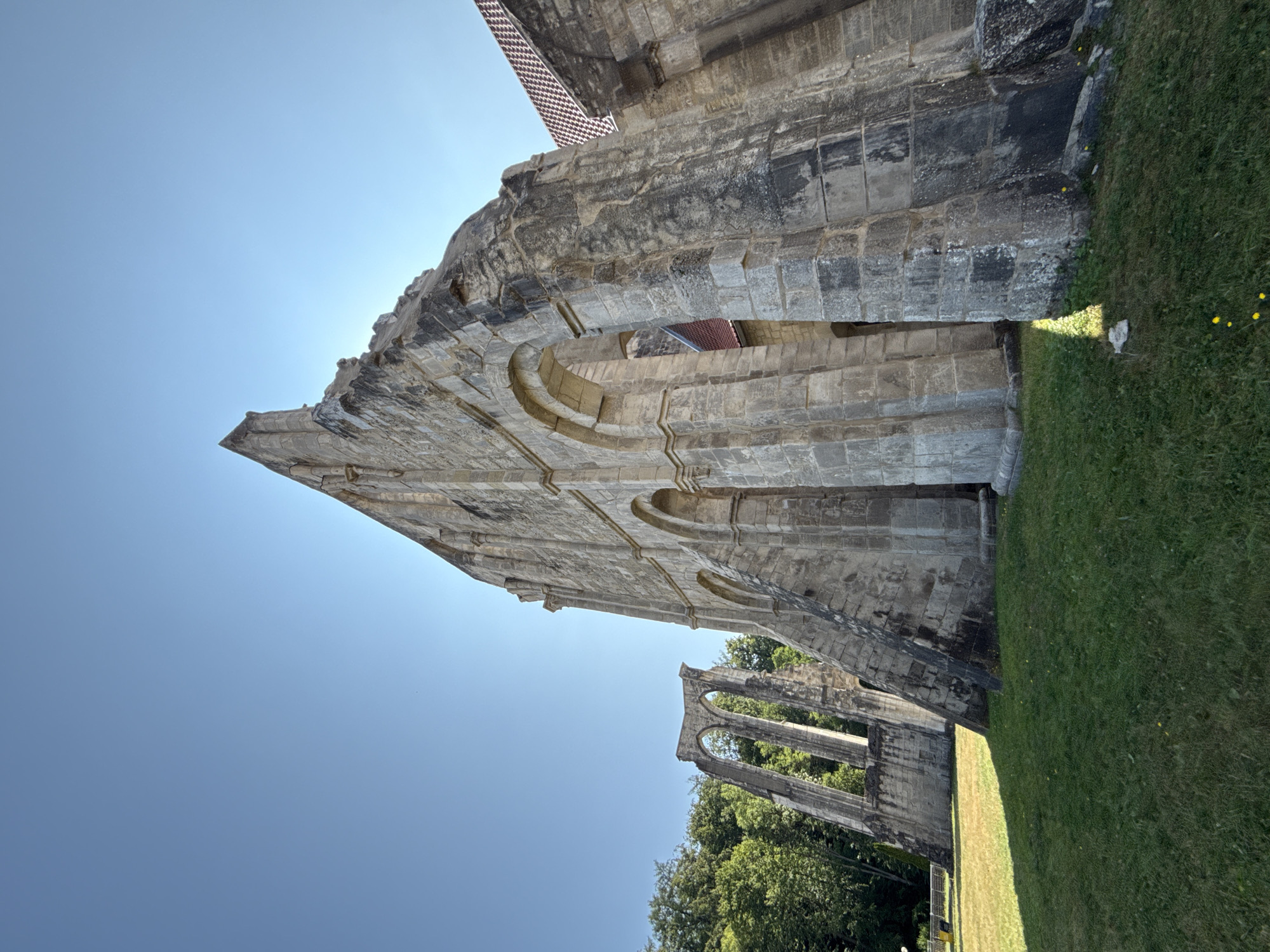 The image shows the exterior ruins of a stone abbey in Walkenried, Germany. The sun is shining brightly, casting shadows on the overgrown grass in front of the structure. The abbey is made of large, weathered stones, with arched openings visible. In the background, lush green trees provide a backdrop against the clear blue sky. The ruins appear to be a historical monument, with no people or animals present in the scene. The overall atmosphere is one of ancient history and natural beauty.
