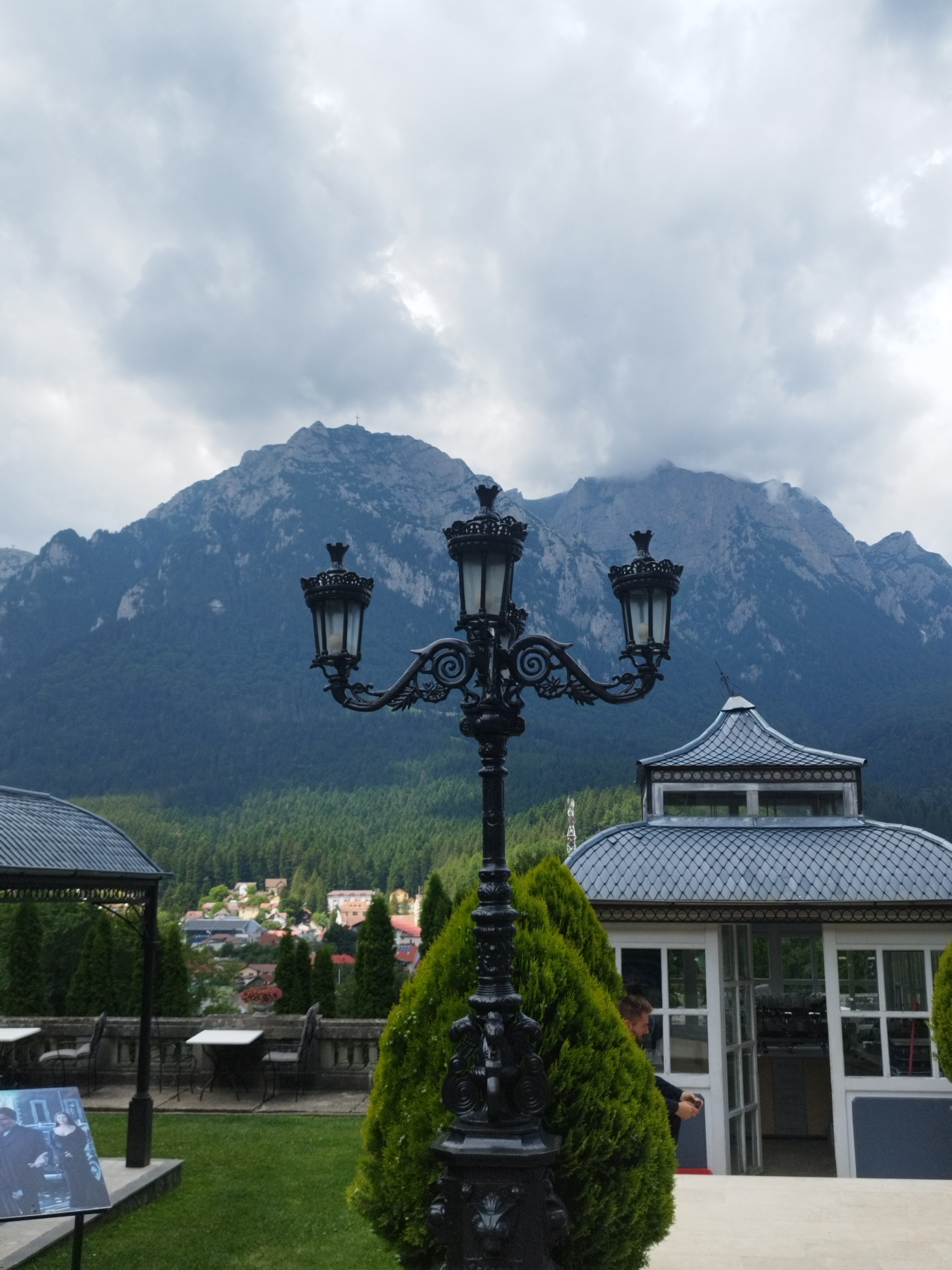 The image captures a view from the terrace of Cantacuzino Castle in Bușteni, Romania, with a prominent ornate lamppost in the foreground. The lamppost is black, featuring three lanterns with intricate detailing, and is positioned on the right side of the frame. To the left of the lamppost, a portion of a gazebo with a dark, corrugated roof is visible. In the mid-ground, there are white tables and chairs arranged on the terrace. Beyond the terrace, a town with colorful rooftops nestles amidst a dense, green forest. Towering over the town and forest is a majestic mountain range under a cloudy sky. The mountain peaks are rugged and partially obscured by clouds, with a cross visible at the summit of one peak. In the far right of the image, a modern glass structure with a grey, tiled roof is visible, and a man is seen exiting through its doorway.