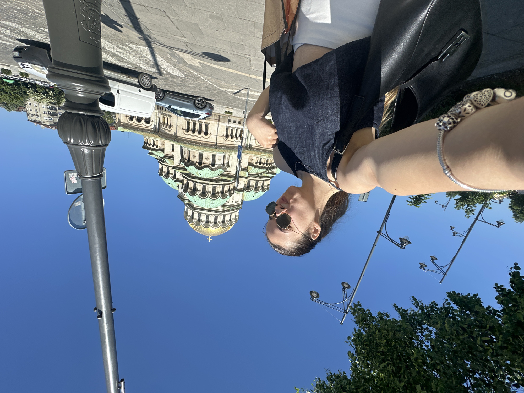 A woman wearing sunglasses, a dark top, and a light skirt takes a selfie in Sofia, Bulgaria. The St. Alexander Nevsky Cathedral, a large Eastern Orthodox cathedral, is visible in the background behind the woman. A street lamp is to the left. Parked cars are also in the background. The sky is a clear blue. A bracelet is seen on the woman's arm.