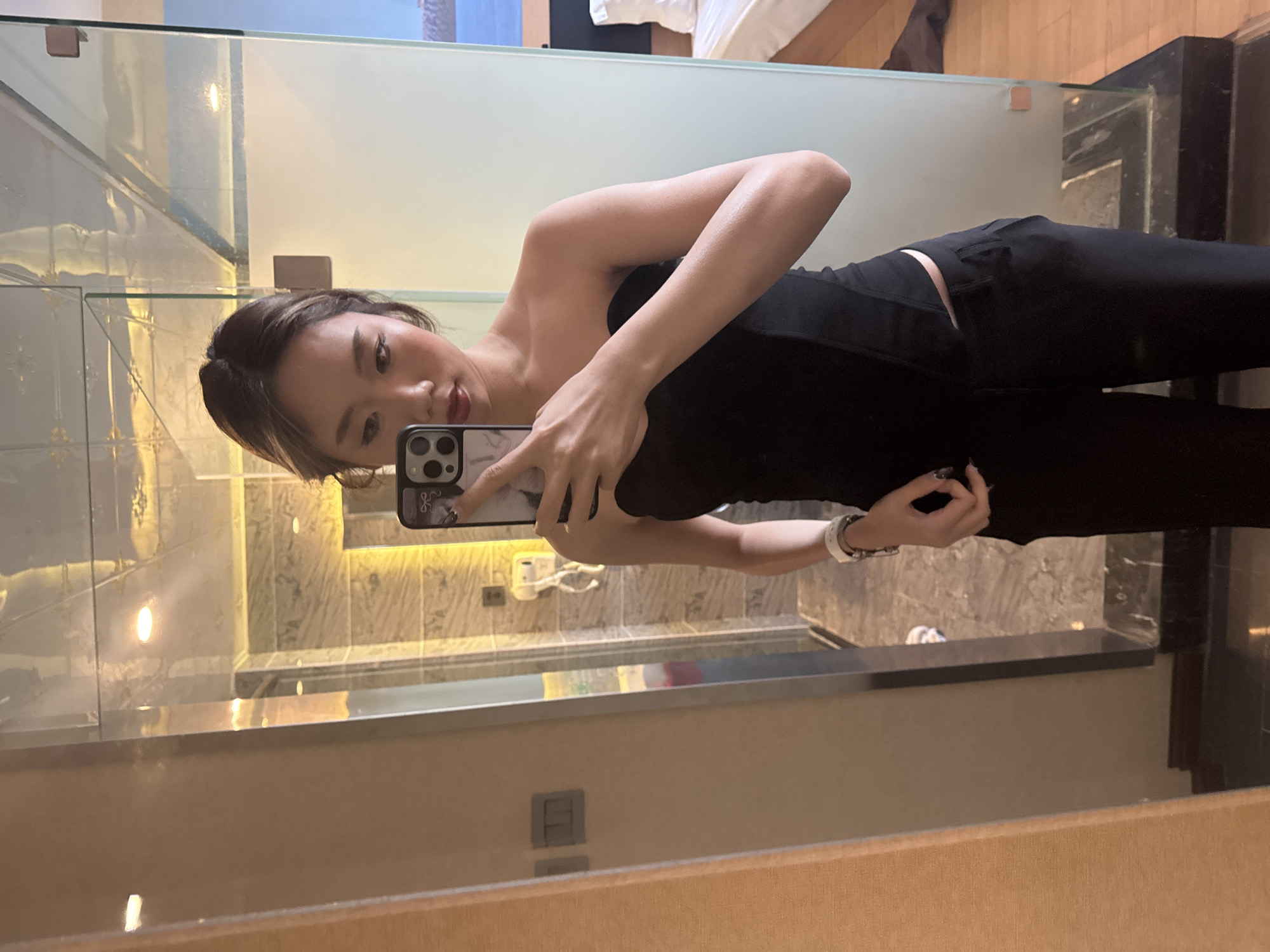 A young East Asian woman is captured in a mirror selfie, standing in what appears to be a modern hotel room or apartment in Ganzhou, China. She is positioned slightly angled to her left, holding a dark-colored smartphone, likely an iPhone, in her right hand to photograph her reflection. The phone features a clear or light-colored case with a distinctive white and dark graphic design on its back.
The woman has shoulder-length dark hair with subtle lighter highlights around her face, and she displays a calm, confident expression. She is dressed in a fitted, strapless black top, possibly corset-style, and dark trousers. A white wristband or watch is visible on her left wrist, and her fingernails are painted a dark color.
The background, visible through the mirror's reflection, showcases a modern interior design. Behind the woman, the mirror reveals a section of a bathroom or dressing area with light-colored, possibly marble or tiled walls, and a dark-colored counter or vanity. A translucent glass partition separates this space from an adjacent room, where a bed with white bedding is partially visible in the upper right corner of the reflection. The scene is illuminated by warm, artificial lighting from recessed fixtures, suggesting an evening or nighttime setting. No discernible text is visible in the image.
