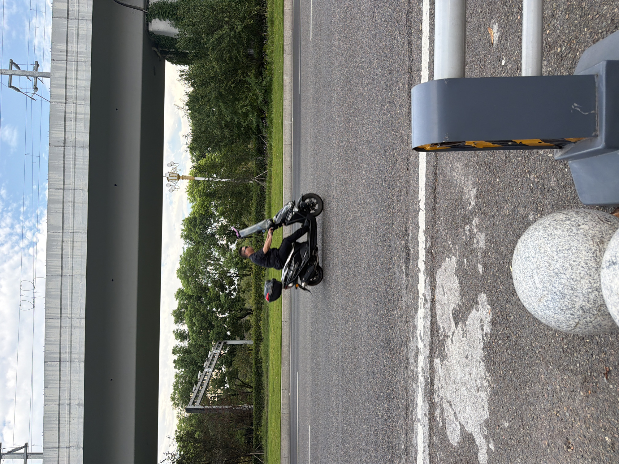 In Jiangjin, China, a man rides a black electric scooter with a red top case on a paved road under an elevated concrete structure. The man is wearing a dark short-sleeved shirt and black pants, with sunglasses. The scooter has a clear windshield. The road has white lane markings. In the background, there are green trees, a light pole with multiple lamps, and power lines above the elevated structure. The foreground shows a section of the road with white markings and a grey bollard with yellow reflective elements.