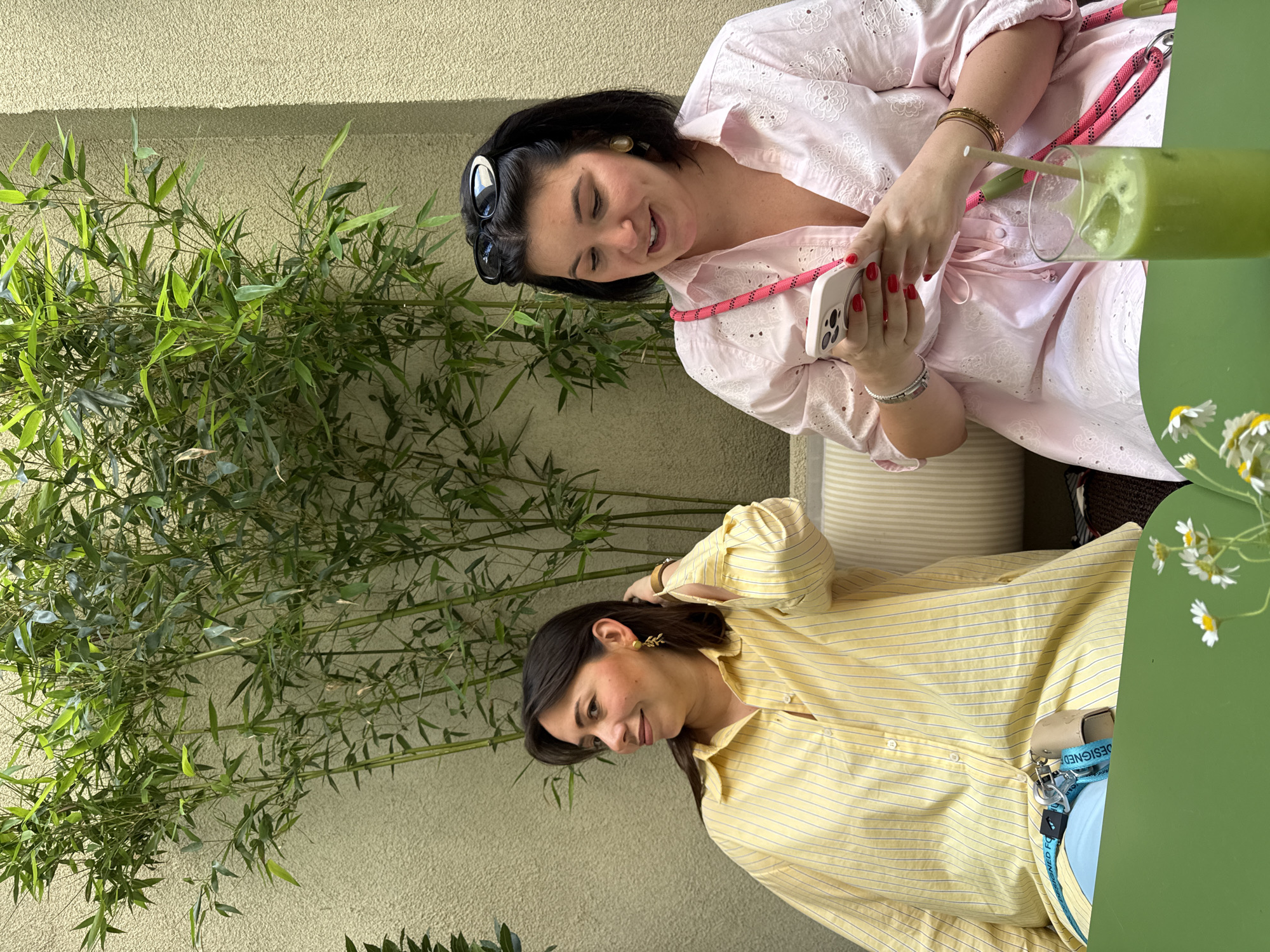 Two women are sitting outside, possibly at a cafe or restaurant in Belgrade, Serbia. The woman on the top is wearing a light pink, eyelet-detailed blouse, has dark hair pulled back with sunglasses perched on her head, and is looking at a smartphone in her hands. She is wearing multiple bracelets and has red nail polish. Next to her is a tall green drink with ice and a straw. The woman on the bottom is wearing a yellow and white striped button-down shirt and has dark hair with gold leaf-shaped earrings. She has her hand on her hair and is looking downwards. Behind them is a wall with bamboo plants. A green table with a vase of daisies is to the right, and a lanyard is visible on the table. They appear relaxed and engaged in a conversation or activity together.
