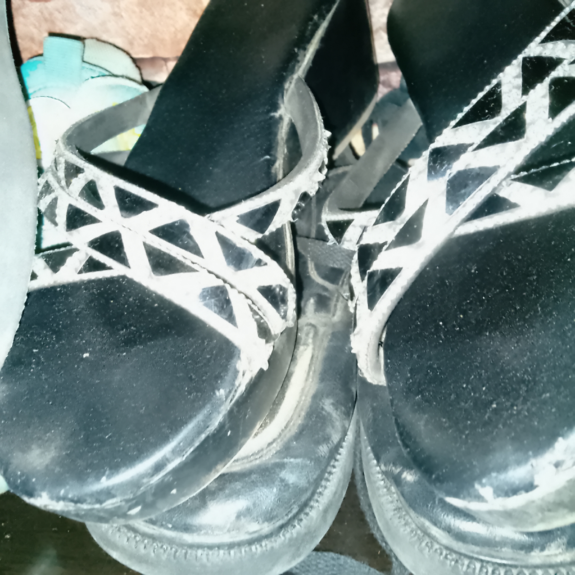 The image shows a close-up of two black platform wedge sandals. The sandals have wide straps with a white geometric pattern. The sole of the left sandal is visible, and it appears to be made of a dark material. The right sandal is partially obscured by the left one. The scene is dimly lit, with some parts of the sandals appearing worn and dusty. There are some other items in the background that are out of focus, including what looks like a light blue and white shoe or sandal on the left. The overall impression is of a candid shot of footwear, possibly stored together. There is no visible text in the image.