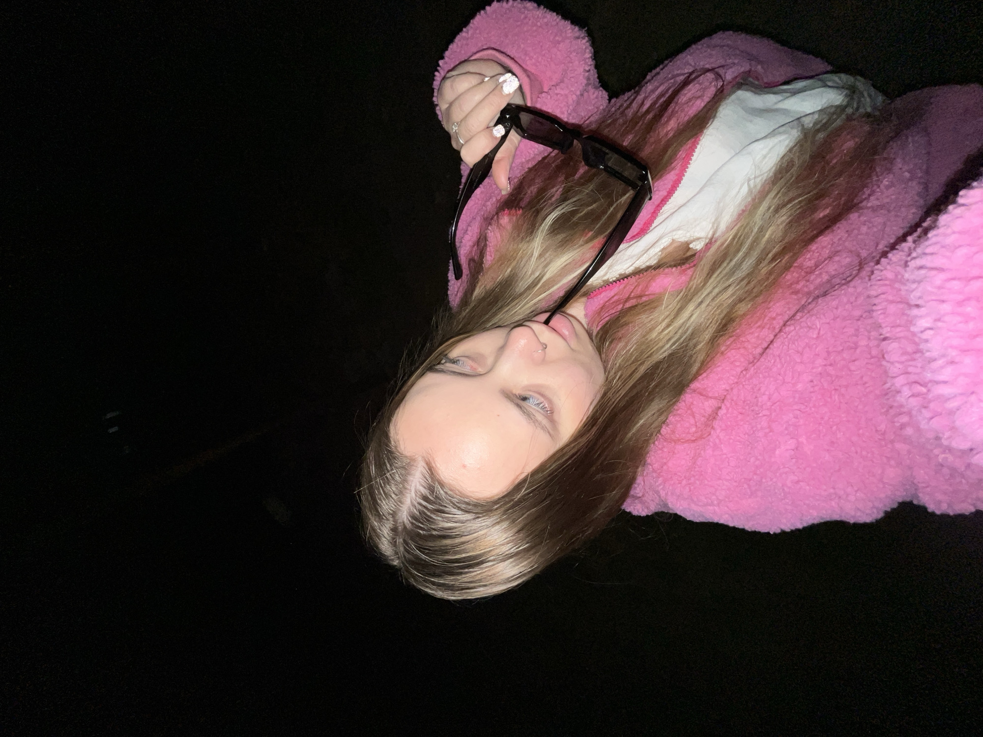 A young woman lies against a dark background. She wears a bright pink, fuzzy jacket over a white shirt. Her blonde hair is parted in the middle. She holds black sunglasses in her right hand, which is adorned with a ring and white nail polish. Her eyes are light-colored. There are no indications of the image location being Novopokrovskaya, Russia.