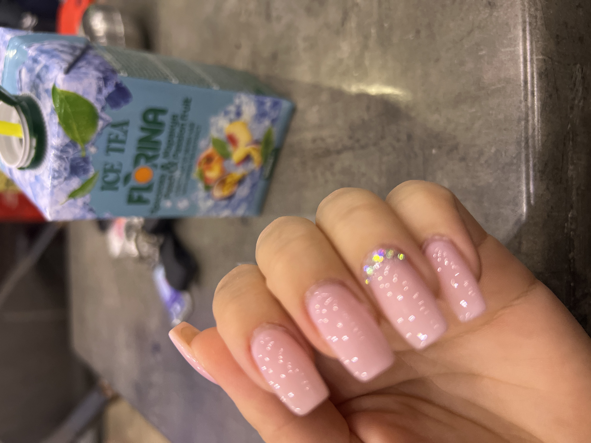 The image shows a person's hand with manicured pink nails. The ring finger has glitter at the tip of the nail. Next to the hand is a carton of "Ice Tea Florina" flavored with peach and passion fruit. The background appears to be a table or counter with a metallic finish. The location is not discernible from the image and cannot be confirmed as Plovdiv, Bulgaria.