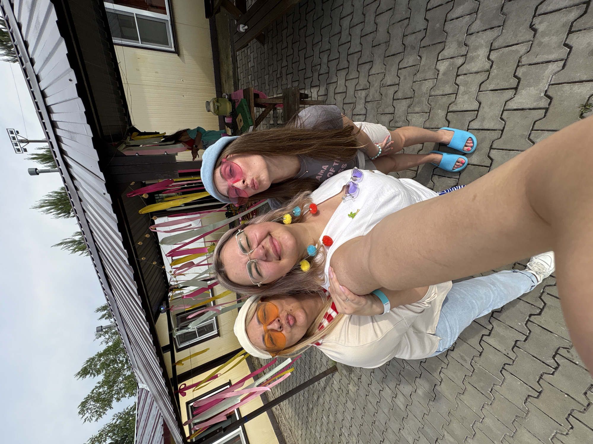 Three women are posing for a selfie outdoors. The woman in the center is wearing glasses and has colorful pom-pom earrings. The woman on the left is wearing orange sunglasses and a white hat. The woman on the right is wearing pink heart-shaped sunglasses and a blue beret. They are standing in front of a building with a corrugated metal roof, decorated with colorful ribbons. The ground is paved with interlocking concrete tiles. In the background, there are trees and a cloudy sky. There is no identifiable information within the image to indicate the location is Birobidzhan, Russia.