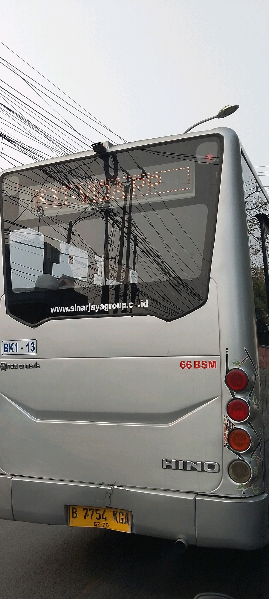 The image shows the rear of a silver bus with an illuminated destination board displaying "BANDUNG KOTA". The bus has a license plate that reads "B 7754 KGA" with "07 . 20" below it. To the right of the license plate, a sticker on the bumper is partially visible. The bus also has "66 BSM" written in red to the right of the destination board. On the left side of the bus, there is a plate with "BK1 - 13" and the "new armada" logo. The bus brand "HINO" is visible on the lower rear. Decorative patterns are present on the right side of the rear of the bus, near the taillights. The scene is set outdoors, with numerous overhead wires crisscrossing the sky, suggesting an urban environment. The location is identified as Bekasi, Indonesia, as indicated by the bus license plate starting with "B".