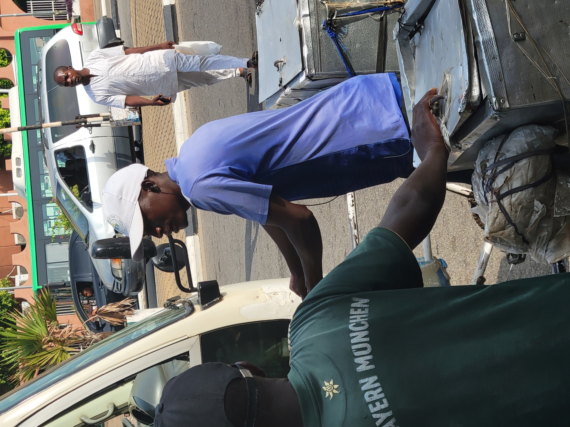 Location: Abuja, Nigeria
The image depicts an outdoor street scene on a sunny day. Several people are visible.
In the foreground, a man wearing a green shirt with the text "BAYERN MUNCHEN" and a black baseball cap is partially visible. Beside him, another man wearing a blue shirt and a white baseball cap is bending over, his arm extended towards a makeshift cart or vehicle.
In the background, a man dressed in white traditional clothing is walking on the sidewalk. Various vehicles, including a white van and a green-striped bus, are parked on the street. The setting appears to be a commercial or residential area with trees and buildings visible along the roadside.
