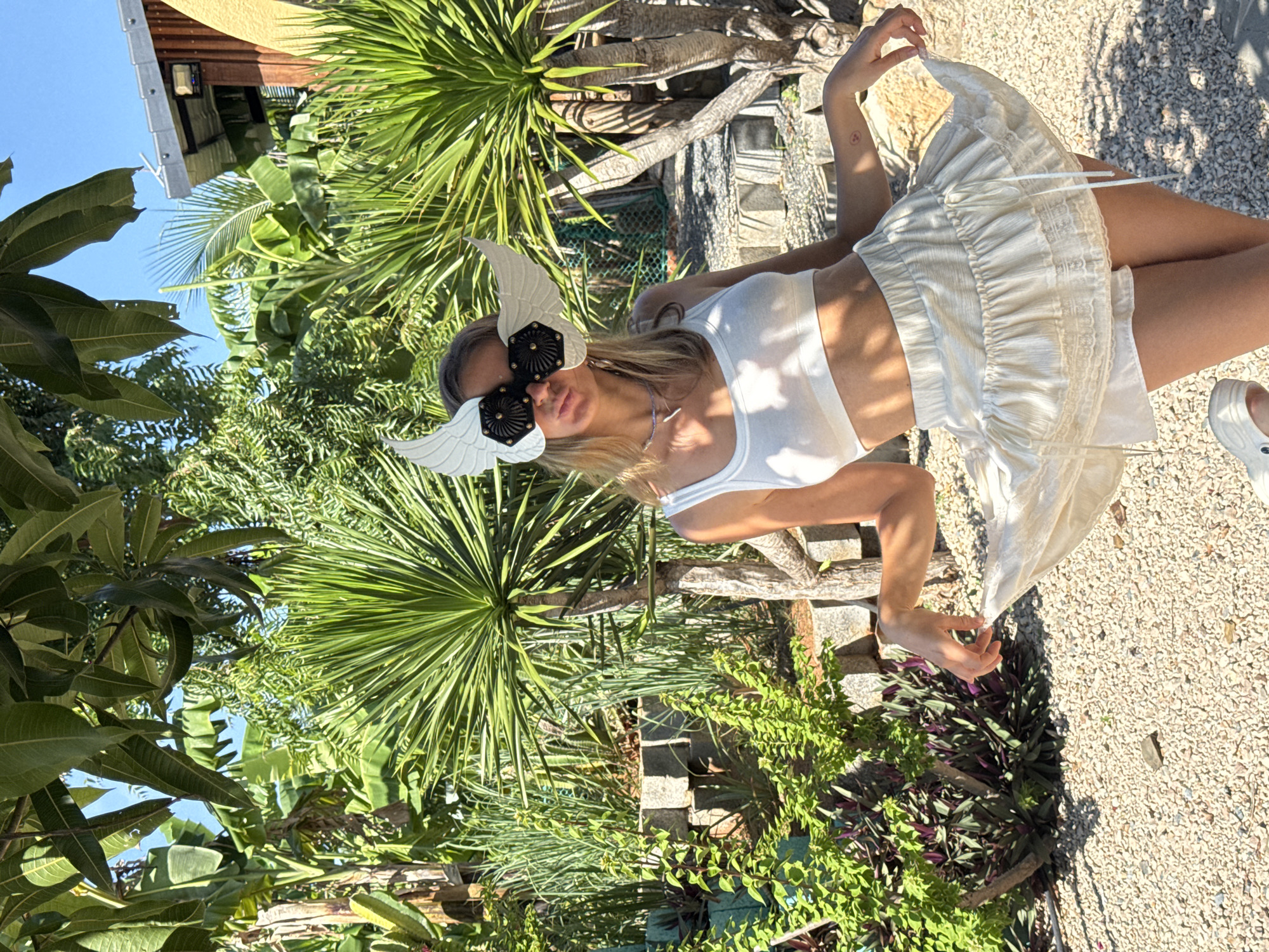 In Mũi Né, Vietnam, a young woman stands in a garden setting. She is wearing a white crop top and a layered, off-white ruffled skirt. Her face is partially obscured by a distinctive mask designed to look like white wings with black decorative elements over the eyes. The background is filled with lush green tropical foliage, including palm trees and other dense vegetation, with a small building visible in the upper right corner. The ground is covered in light-colored gravel. The woman is posed with one hand lightly holding the edge of her skirt, creating a slight movement effect.