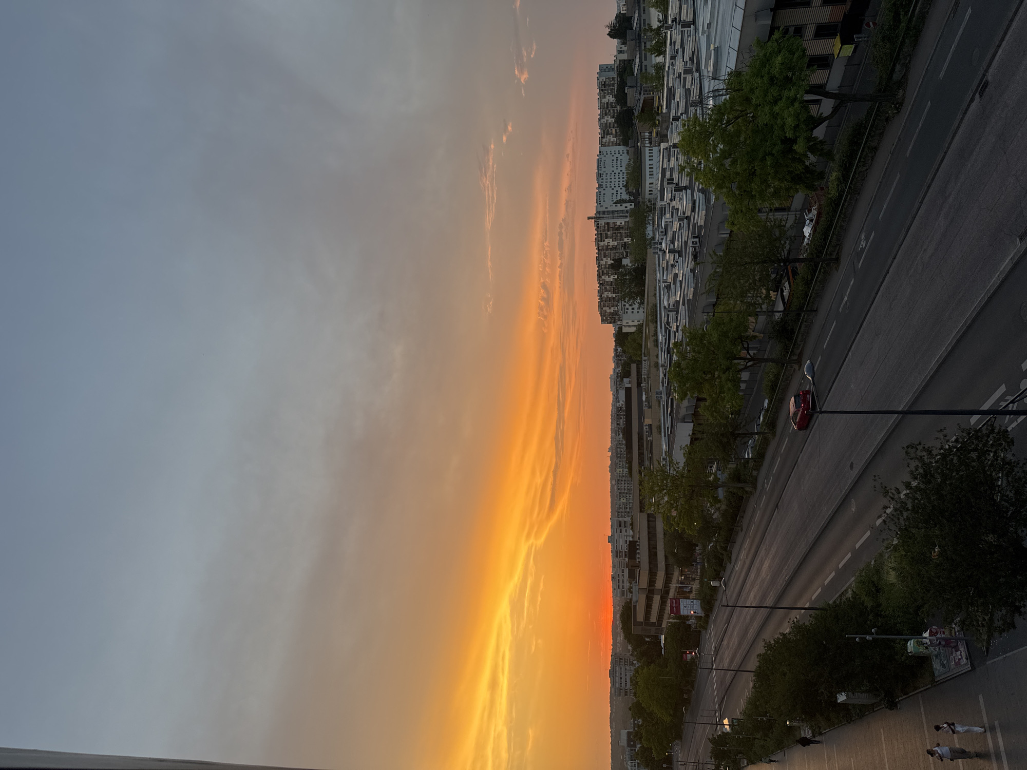 The image shows a wide view of a city at sunset. The sky is a vibrant orange and yellow on the left, transitioning to a lighter blue and gray on the right, with wispy clouds. In the foreground, a street runs diagonally across the frame, bordered by trees and sidewalks. Buildings, some appearing to be residential and others industrial with flat, white roofs covered in air conditioning units, line the street and the background. There are a few cars on the street, including a red one. On the sidewalk on the left, two individuals are walking. The overall scene depicts an urban environment during the golden hour.