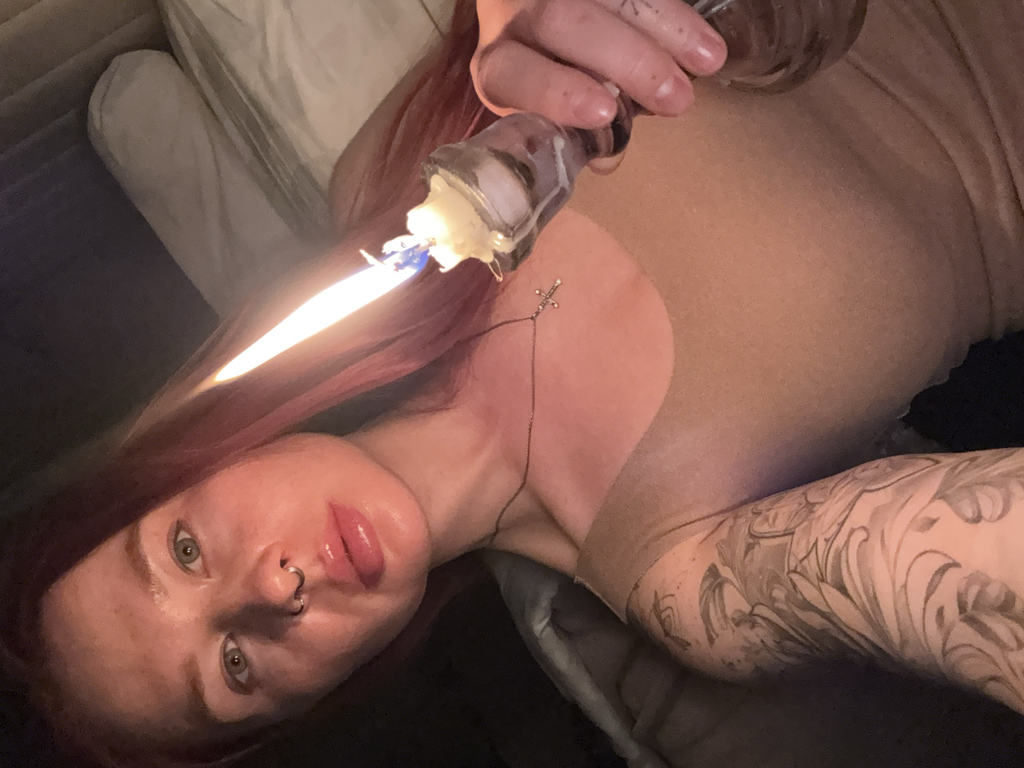 An indoor, close-up shot features a young adult, likely female, with red hair and light skin, lying down and looking towards the camera. She has green eyes, a septum piercing, and a small cross pendant on a necklace. Her left arm is adorned with a prominent tattoo featuring an intricate, scrolling pattern. She is wearing a light-colored, possibly nude or sheer, garment over her torso.
She holds a clear glass object, resembling a small bottle or candle holder, in her right hand. A lit candle is inserted into this object, emitting a bright, elongated flame that illuminates her face and the immediate surroundings. The overall lighting is dim, suggesting it is evening or night. The background shows pillows and blankets, indicating she is on a bed or sofa. Her expression is direct and somewhat neutral, with her lips appearing glossy. No specific location cues are visible within the image, although external context indicates Rostov-on-Don, Russia. There is no visible text.