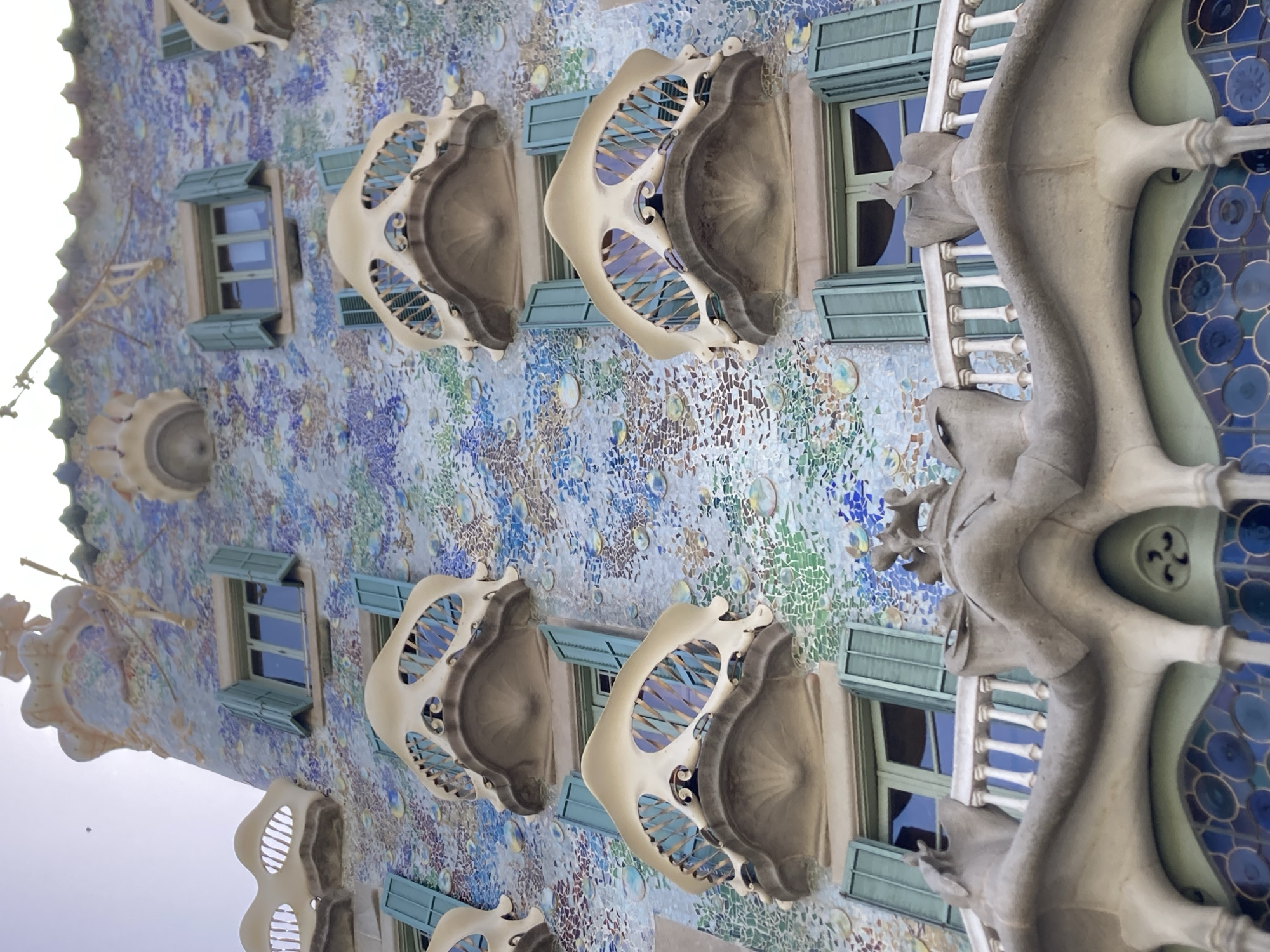The image displays the exterior of a building with a highly ornate and colorful facade. The building appears to be a work of art nouveau architecture, with irregular shapes, vibrant mosaics, and whimsical decorative elements. Several windows with light green shutters are visible. Some windows are framed by sculptural, bone-like balconies that project outwards. The walls are covered in a mosaic of small, irregular tiles in various shades of blue, green, yellow, and purple, creating a flowing, organic pattern. Larger circular decorative elements, some with intricate patterns, are also integrated into the facade.
The perspective is angled, making it difficult to determine the exact orientation of the building, but it seems to be a close-up view of a section of the facade. There are no people or explicit activities taking place in the image. The lighting suggests it is daytime, possibly sunny, given the brightness and reflections on the windows. The overall impression is one of elaborate craftsmanship and artistic expression. The unique architectural style strongly suggests it is Casa Batlló in Barcelona, Spain, a famous landmark designed by Antoni Gaudí, known for its organic forms and vibrant tile work. There is no visible text in the image.