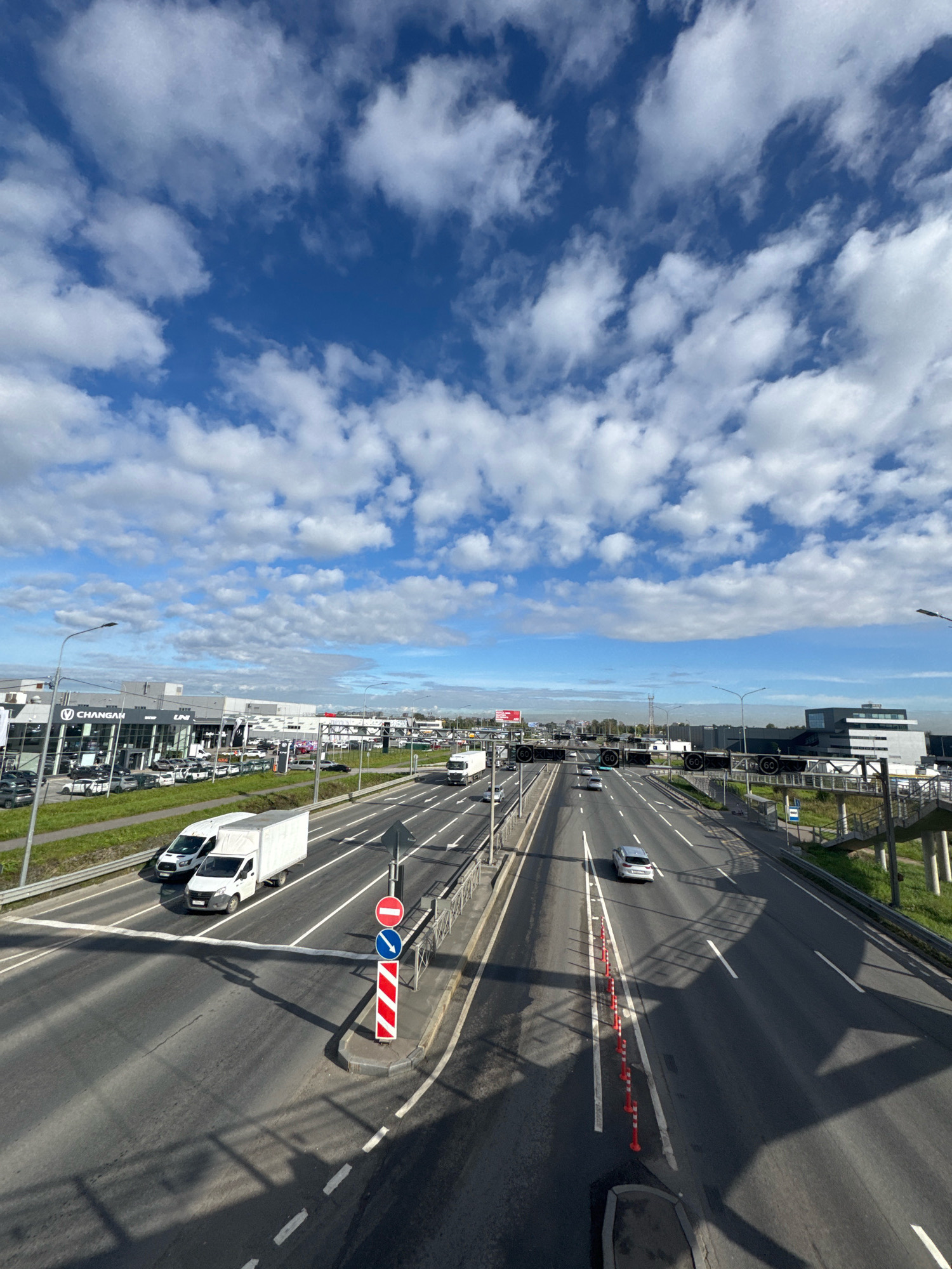 This is a vertical, eye-level, wide shot of a multi-lane highway in Saint Petersburg, Russia. The sky is bright blue with scattered white clouds. The highway is busy with vehicles including trucks, vans, and cars. On the left side of the highway, there is a car dealership with several vehicles on display. In the distance, there are several industrial buildings. The road is well-maintained with clear lane markings and traffic signs. The overall impression is one of a modern, active urban environment on a clear day.