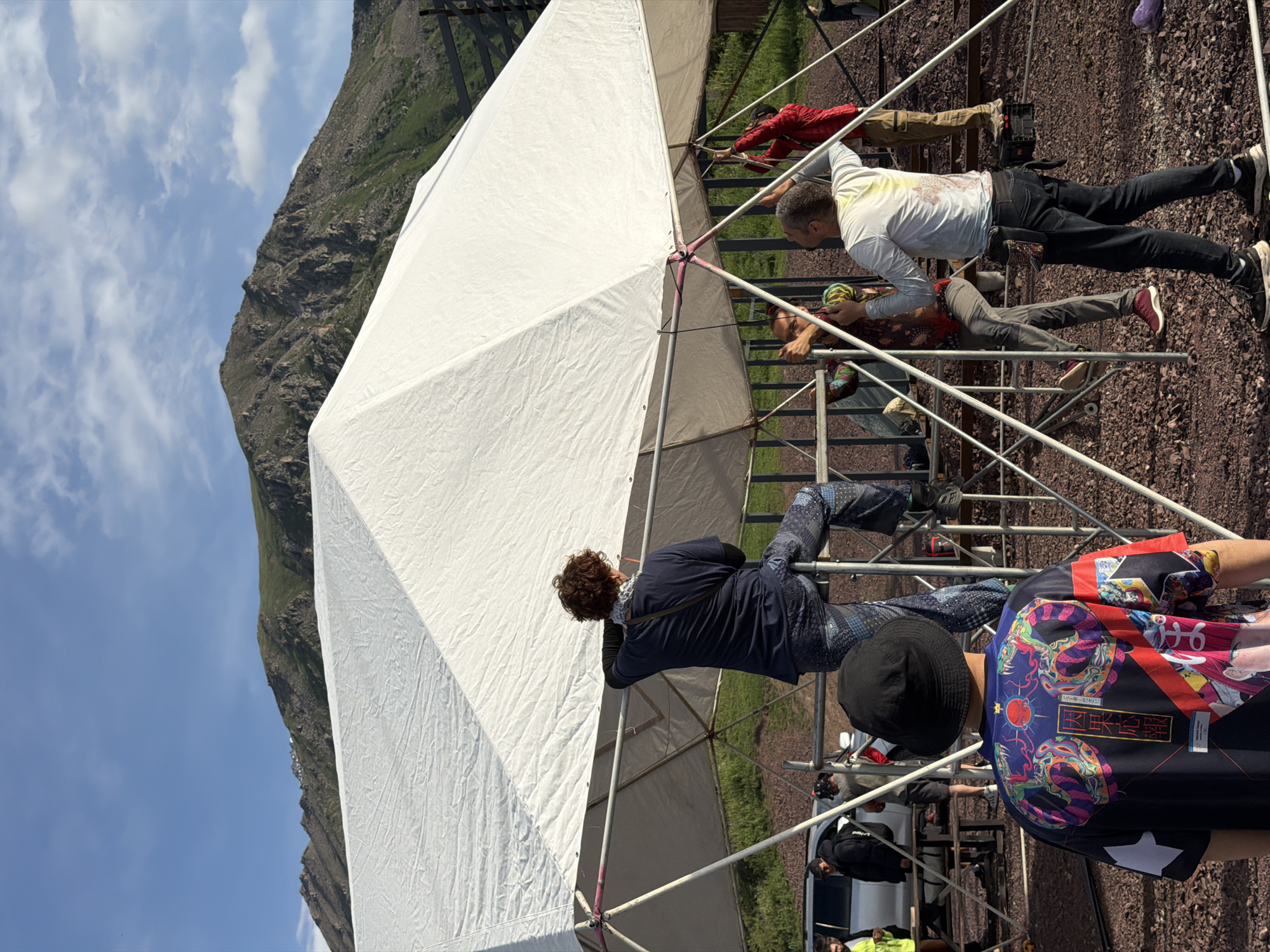 The image shows a group of people gathered around a large, white, tent-like structure supported by a metal frame. The scene appears to be outdoors, with a rocky, green mountain and a bright blue sky with some clouds in the background. The ground is covered in gravel. Several individuals are visible on and around the structure, some climbing on it and others observing. One person in a red jacket is reaching up to the structure. Another person in a white t-shirt and dark pants is leaning over. A person in a dark blue top and patterned pants is holding onto the frame. Towards the bottom right, a person in a colorful, patterned shirt and a black bucket hat is visible. The people's clothing suggests a casual, outdoor event. The time of day is likely daytime, given the bright sunlight and clear sky. The location cues, such as the mountain landscape and the gravel ground, are consistent with the provided context of Kashka Suu, Kyrgyzstan. There is visible text on the colorful shirt, which appears to be in Japanese characters and also features graphic designs of skulls and other imagery. The overall impression is of an outdoor gathering or event in a mountainous region.