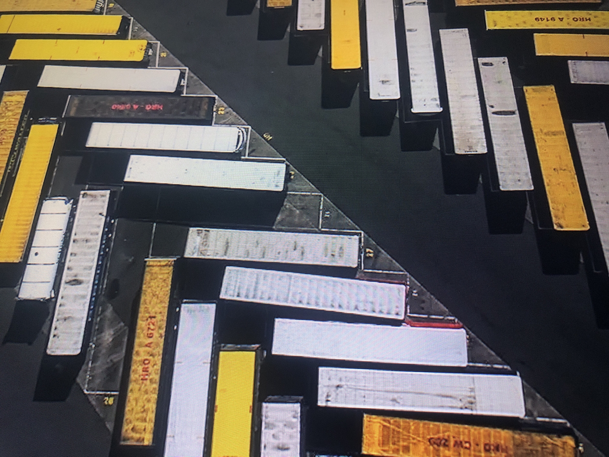 The image depicts an aerial view of a shipping container yard. The yard is divided by a dark, linear pathway or boundary. Containers are arranged in neat rows on both sides of this division. The containers vary in color, primarily yellow, white, and gray. Some containers have markings or labels, including "HRO A 9149" and "HRO A 6721" visible on some. The environment is industrial, with the containers densely packed and organized. The location is likely in a port area within the city of Buenos Aires, Argentina, given the presence of shipping containers and the location on the map.