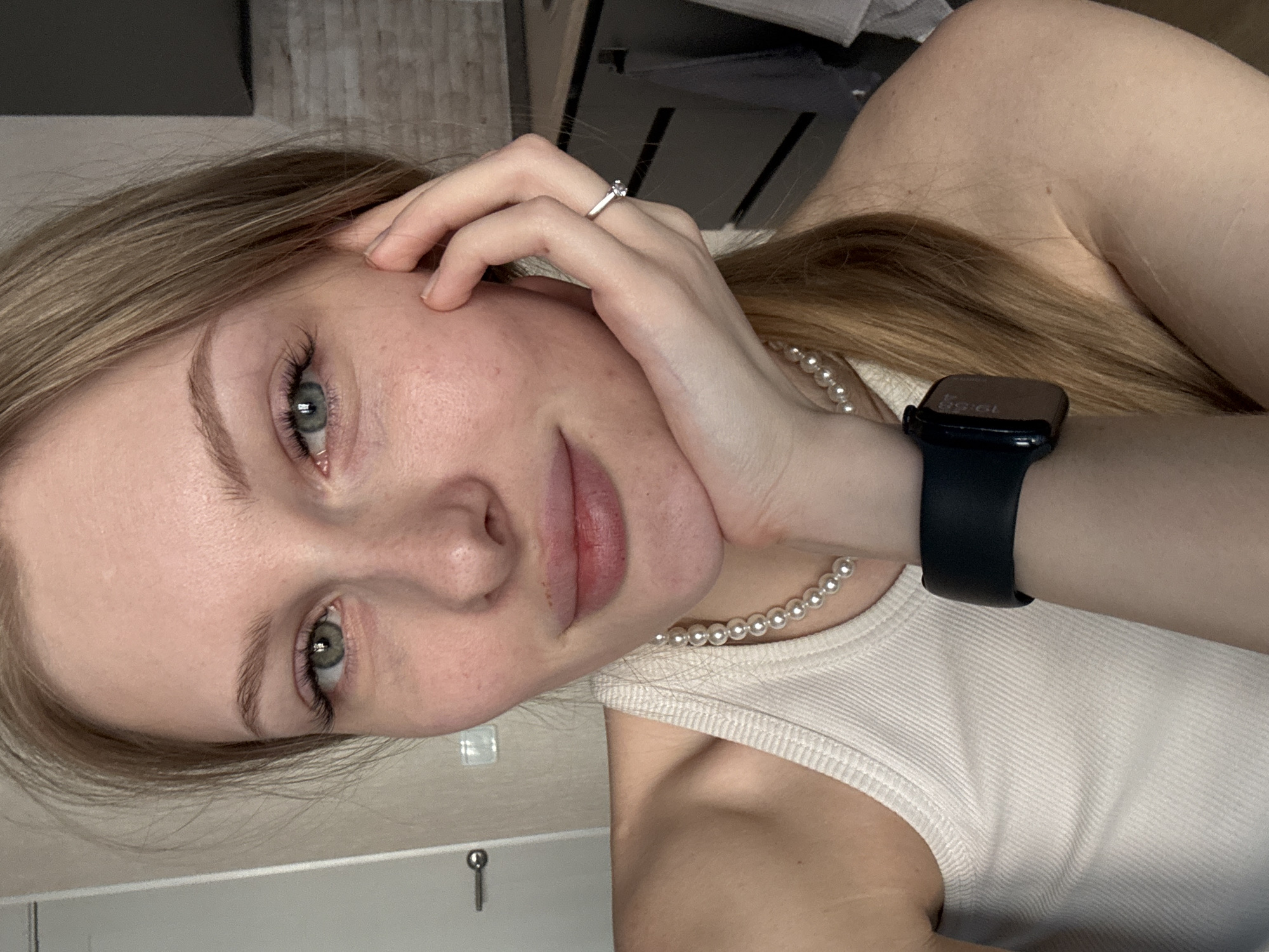 A close-up selfie shows a young woman indoors in Perm, Russia. She has light blonde or light brown hair that falls around her face and over her shoulder, and striking blue-green eyes. Her skin appears fair. She is looking directly at the camera with a neutral to slight smile. Her left hand is raised, with fingers gently resting against her cheek and forehead. On her ring finger, she wears a silver ring with a single small, clear stone.
She is wearing a simple white or off-white ribbed tank top. Around her neck, a delicate pearl necklace is visible. On her left wrist, she wears a black smartwatch, which displays the time "19:58". The background is dimly lit and somewhat out of focus, suggesting an indoor setting with walls and possibly some furniture. The image is oriented diagonally, as if taken while lying down or leaning back.