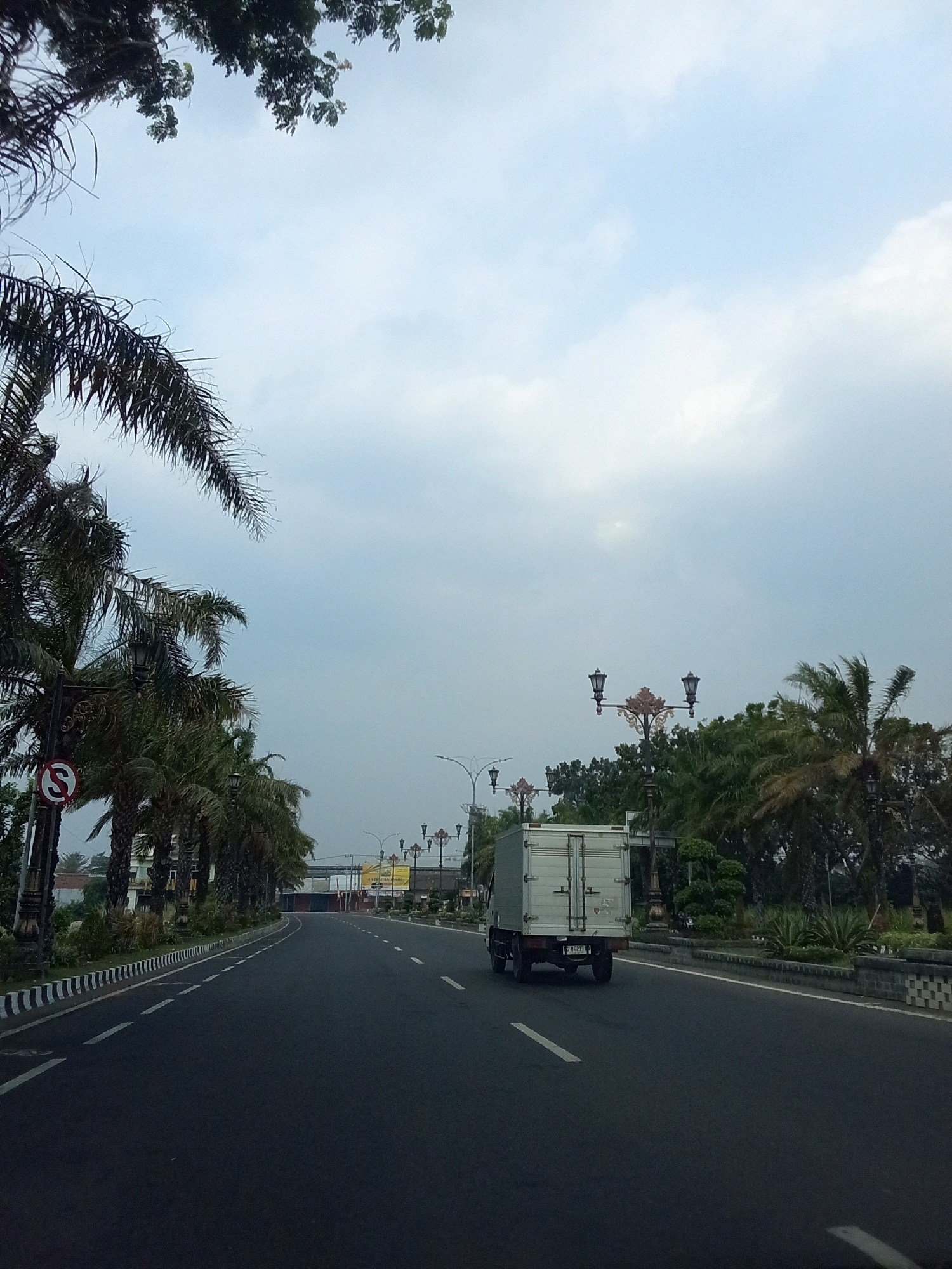 The image depicts a two-lane asphalt road in Gurah, Indonesia, with a white truck driving away from the viewer in the right lane. The road is bordered by a sidewalk with black and white striped markings on the left and a stone wall with greenery on the right. Palm trees line the left side of the road, with a "No Stopping" sign visible on a pole. Ornate street lamps are present on both sides of the road, with more trees and buildings visible in the background. The sky is overcast with scattered clouds. The overall scene suggests a somewhat overcast day in a semi-urban or suburban area.