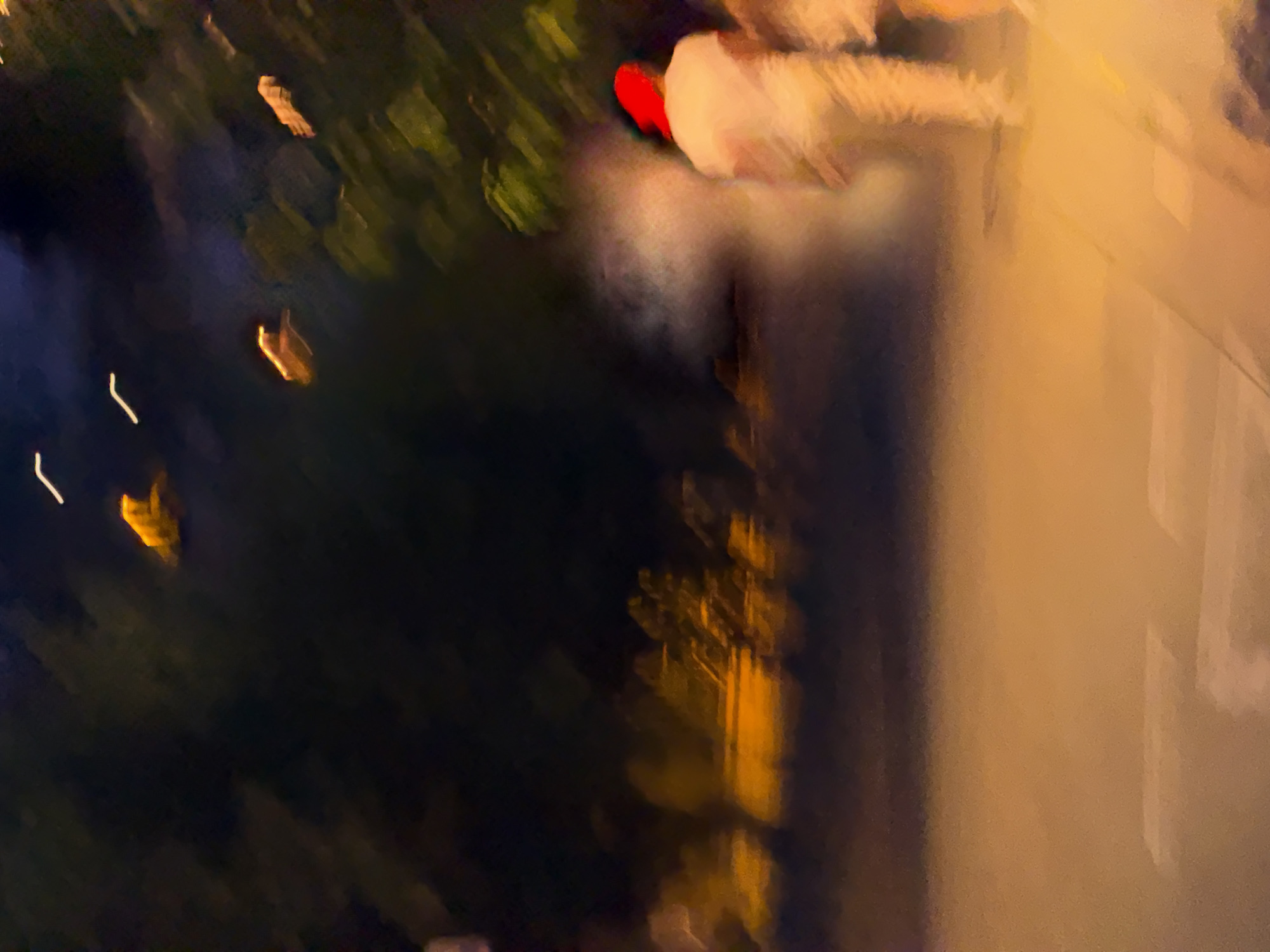 The image depicts a nighttime scene, possibly in Blanes, Spain, with a blurred and impressionistic quality. In the foreground, a person wearing a white shirt and a red head covering is visible, walking away from the camera. They are partially obscured by a hazy, white mist or smoke. Behind them, the environment is dark with streaks of light that suggest movement, possibly from passing vehicles or fireworks. Trees are visible in the background, their forms softened by the low light and motion blur. The ground appears to be a paved surface, with illuminated areas suggesting streetlights or nearby buildings. Several indistinct, bright orange and yellow shapes are scattered throughout the upper and middle portions of the image, which could be distant lights or abstract representations of illuminated objects. The overall impression is one of a dynamic and somewhat mysterious urban night.