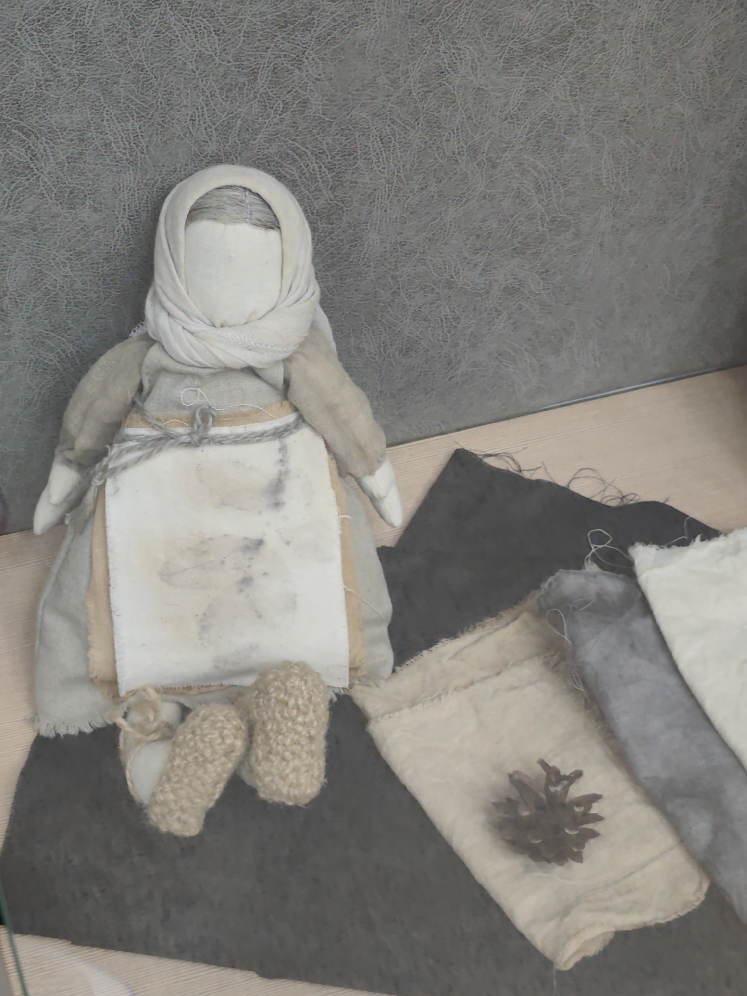 A handmade doll, dressed in traditional attire with a headscarf, apron, and long dress, sits on a surface. The doll's face is featureless, and its hands are white. The doll has knitted booties on its feet. Around the doll, there are several pieces of fabric in various shades of gray and off-white. A pinecone rests on one of the fabric pieces. The background is a textured gray wall. The scene appears to be an exhibit or display, possibly in a museum or cultural center. The doll's construction and the accompanying materials suggest a focus on traditional crafts or folk culture, with a potential link to the natural environment.