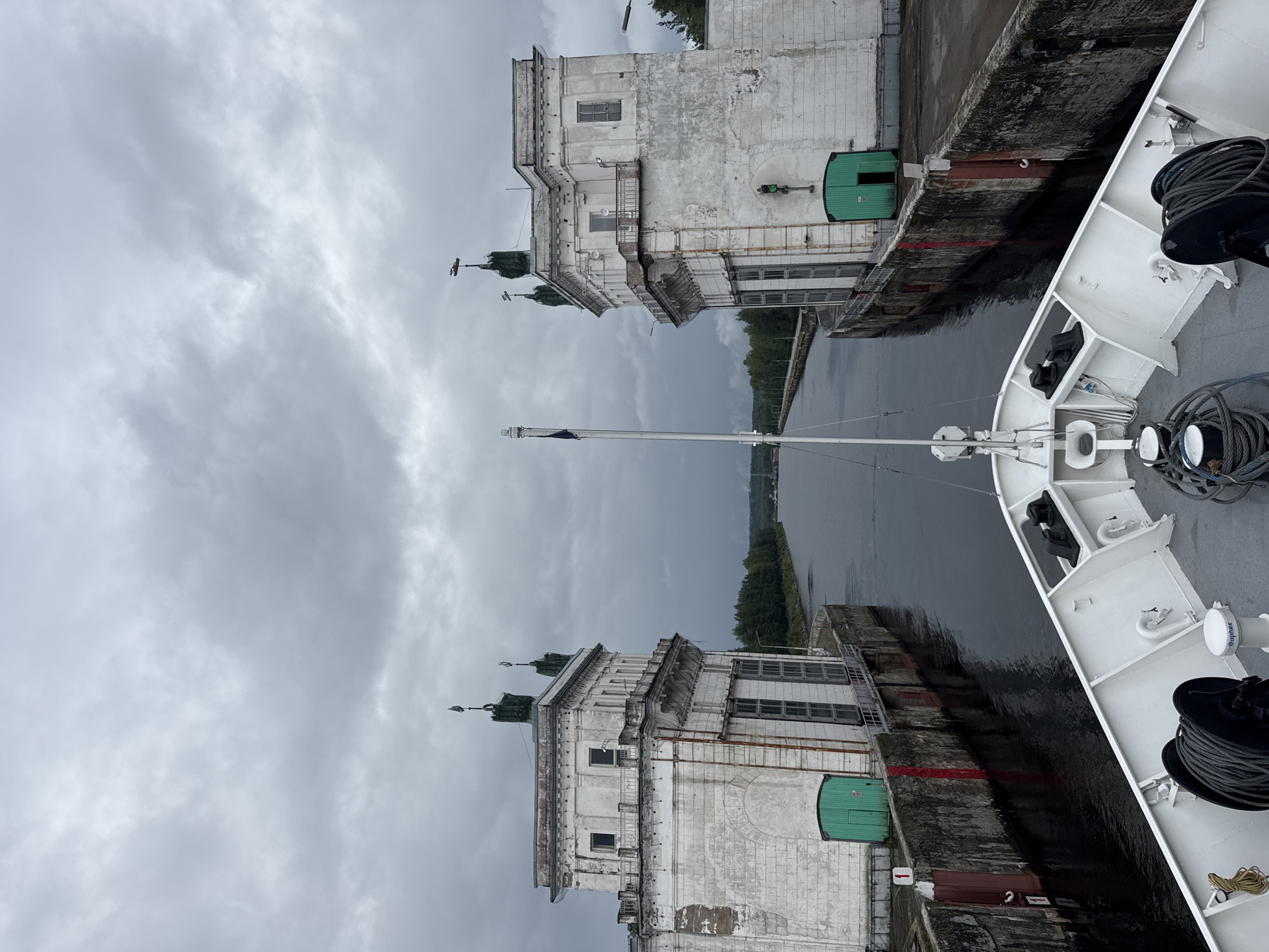 The image shows a view from the bow of a white ship as it approaches a canal lock. Two imposing, weathered white buildings flank the canal, with green doors visible on each. A flag flies from a tall pole on the left side of the image. The sky is heavily overcast with dark clouds. The canal itself is dark and reflects the dull sky. The ship's deck is visible in the foreground, featuring mooring ropes and winches. The context suggests this is the Verkhne-Svirsky Lock, part of the V.I. Lenin Volga-Don Canal system, located near Podporozhye, Russia, a significant waterway for navigation in the region.