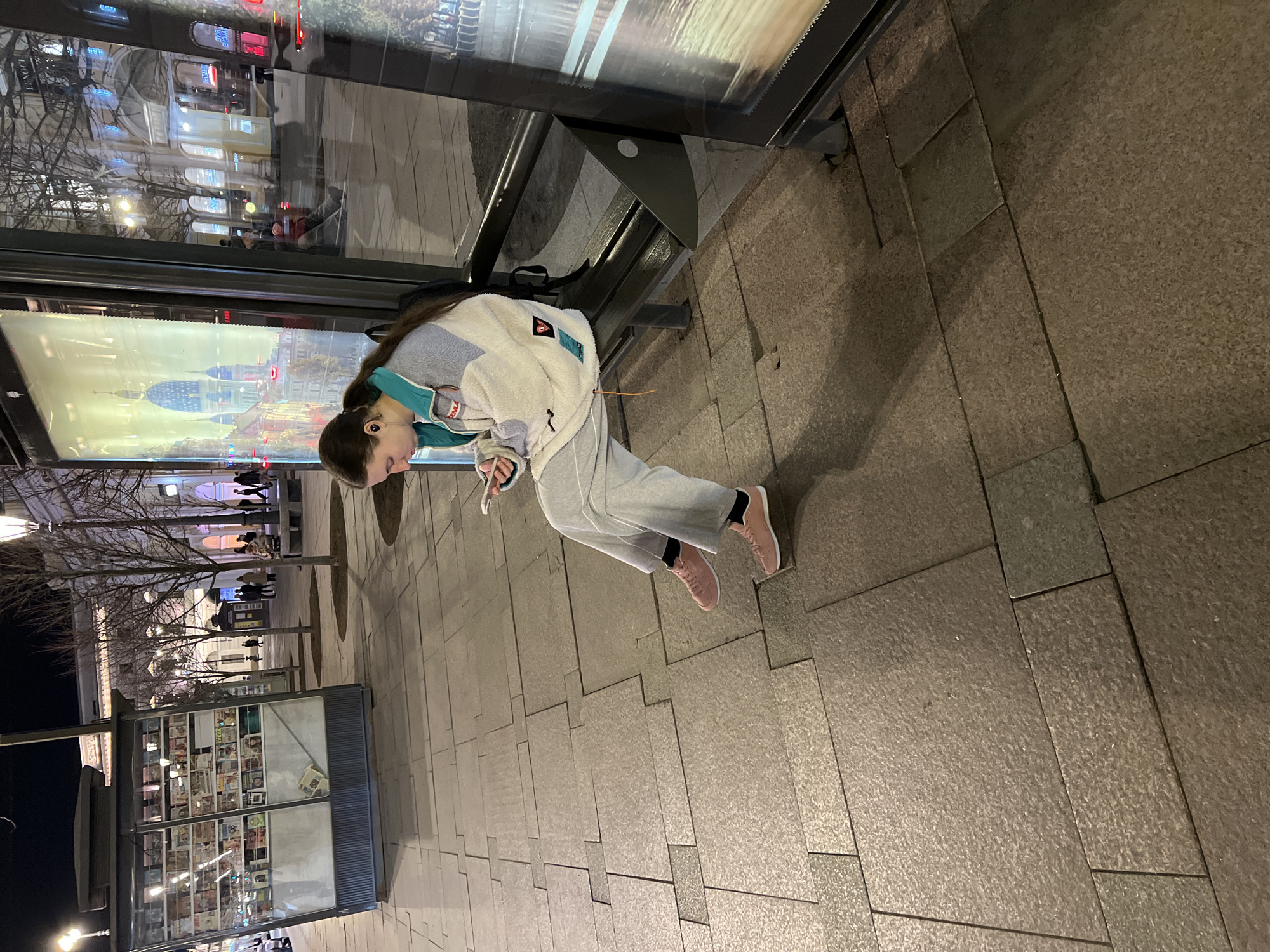 A young woman in a white and gray jacket and gray sweatpants, wearing pink sneakers, sits on a bench at a bus stop in Saint Petersburg, Russia. She is looking at her phone. The bus stop has a large digital display screen showing a picture of a building with a large blue dome. A newsstand is nearby, filled with various publications. Bare trees line the street. The ground is paved with stone blocks.