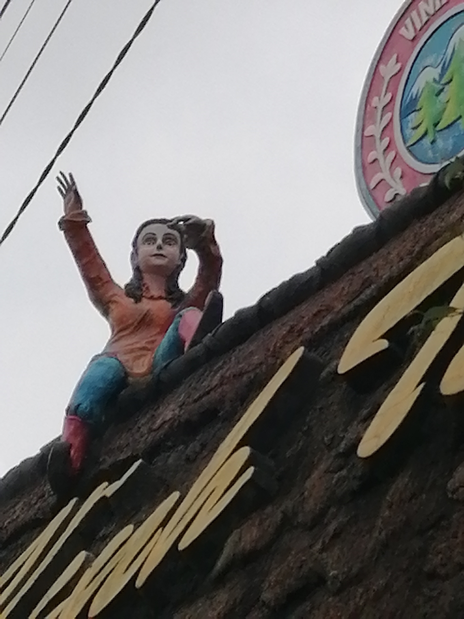 A medium shot captures a painted statue of a young woman perched on a dark, textured surface, likely a building facade, against an overcast sky. The statue, depicting a person with dark hair, wears an orange long-sleeved top, blue trousers, and pink or red footwear. She is seated with her left arm raised, hand open, and her right hand near her head, looking upwards.
To the right of the statue, a circular sign is partially visible. It has a pink outer ring with white floral or leaf patterns, encircling a blue center adorned with green pine trees and white elements, possibly stylized mountains or clouds. The letters "VIN" are visible at the top of this sign. Below the statue and extending across the textured surface are large, light-colored, stylized letters, partially obscured, which appear to be part of a business name or decorative element. Thin, dark power lines stretch across the light grey sky in the upper left of the frame. The overall impression is an outdoor display, possibly from a shop or attraction in Cam Ly - Đà Lạt, Vietnam, with elements that evoke the region's characteristic pine forests. The lighting suggests daylight under a cloudy sky.
