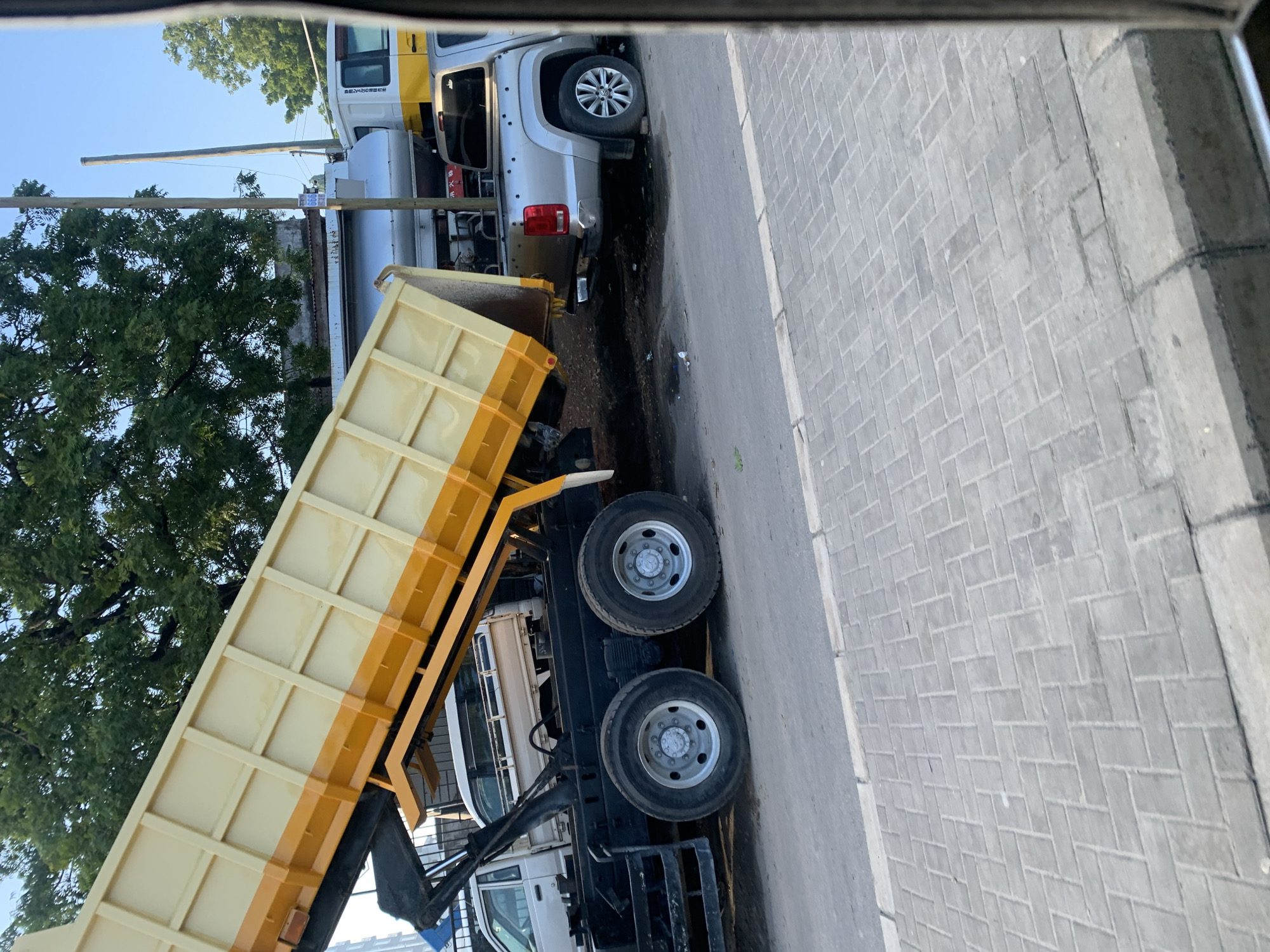 In the city of Dar es Salaam, Tanzania, a light yellow dump truck with a yellow and orange bed is parked on a paved road. The truck's bed is raised at an angle, suggesting it is either dumping or being loaded. The truck has dual rear wheels. In front of the dump truck is a silver pickup truck. To the left of the dump truck, a white truck is partially visible. In the background, there are trees, buildings, and utility poles. The sky is clear and blue. The scene appears to be an outdoor industrial or service area.