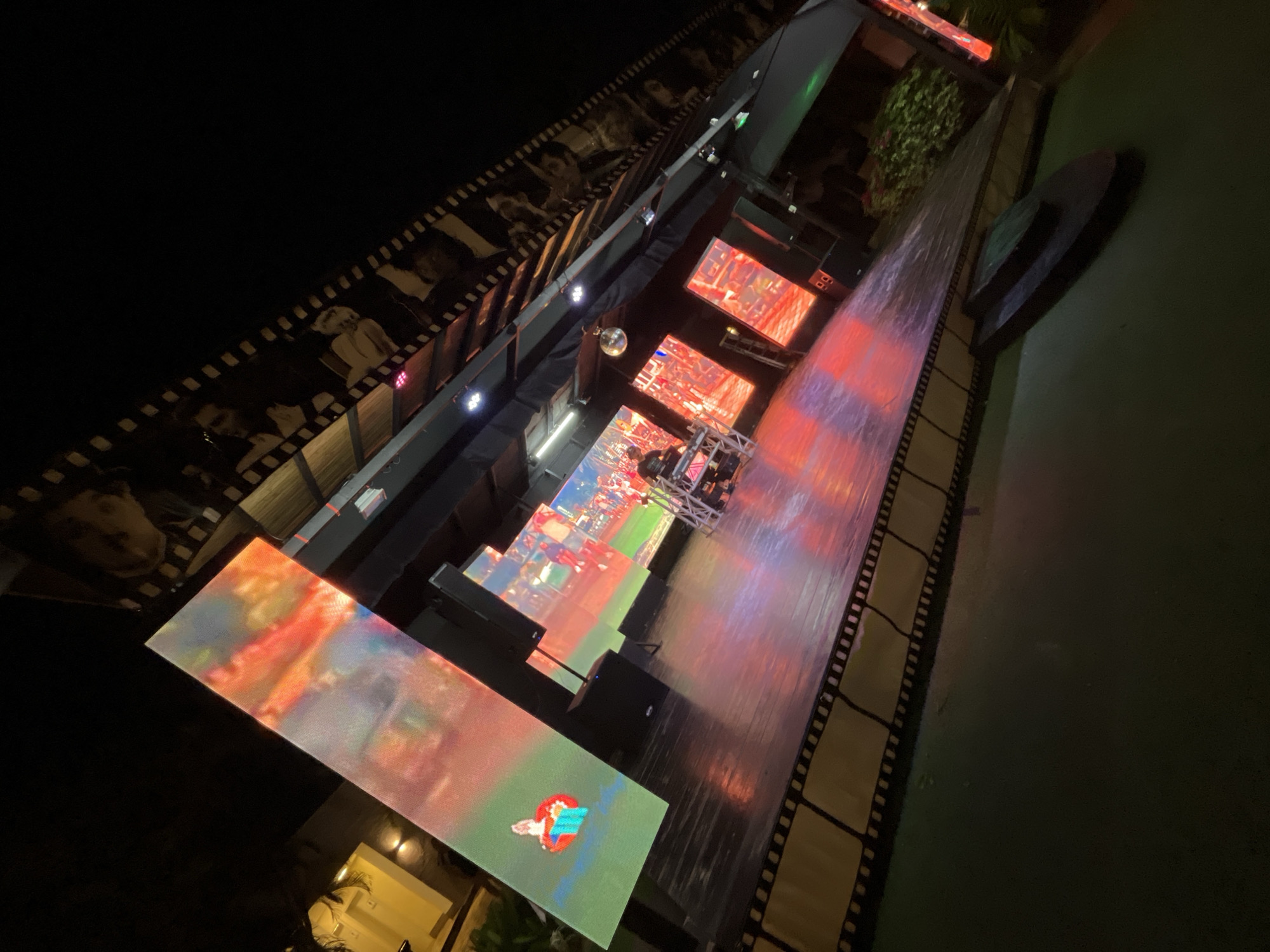 At night, the image shows a walkway with large video screens displaying colorful images and a filmstrip motif lining the edges. Above, there is a structure with portraits along its side and a disco ball is visible. It appears to be a public space or event location. The city is Porlamar, Venezuela.