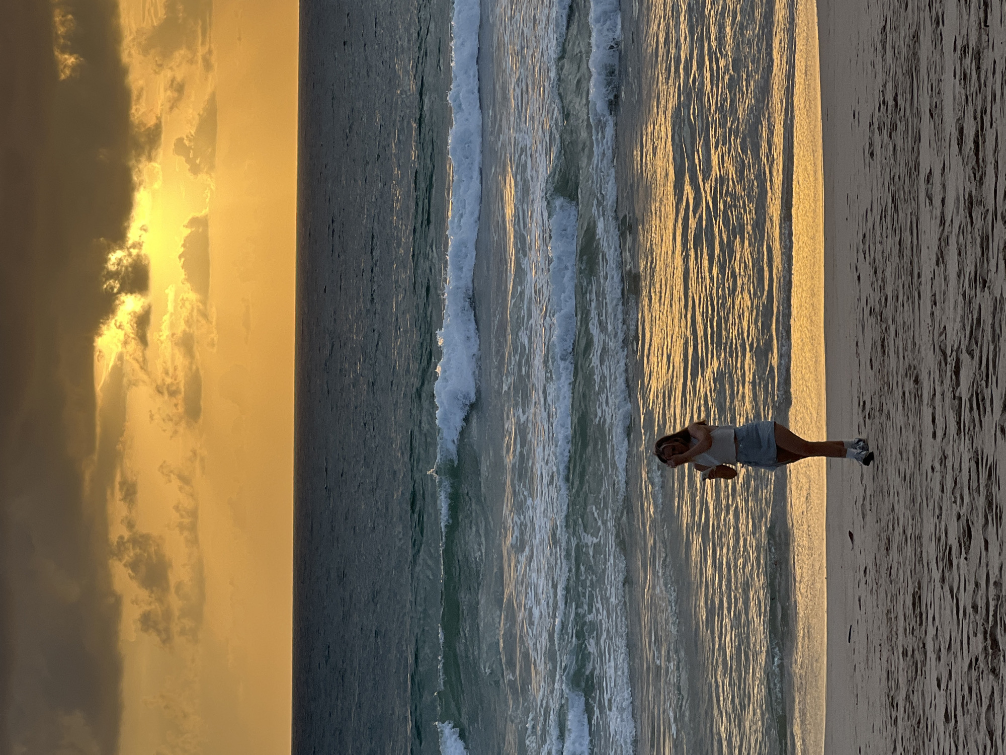 A full-shot, vertical, outdoor, natural light photograph captures a young woman on a beach during sunset. The woman, with long brown hair and wearing a white top and denim skirt, is standing on the sand near the water's edge, holding a phone in her right hand and touching her face with her left. The ocean is in the background with multiple rows of waves breaking. The sky above is filled with dark clouds illuminated by a bright orange sunset. The location is the beach in Choeng Thale, Thailand.