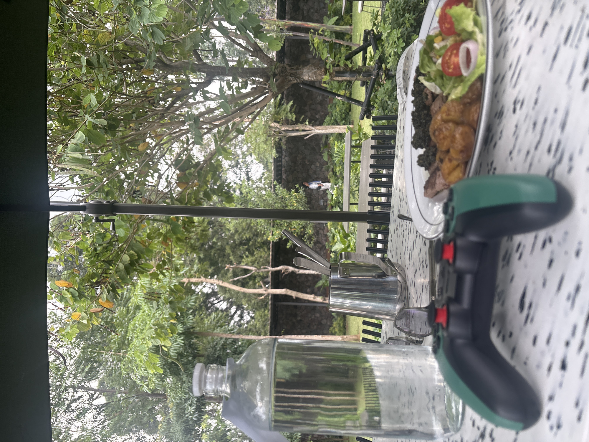 The image depicts a table set for a meal outdoors, likely at a cafe or restaurant in Bandung City, Indonesia. In the foreground, a video game controller with black and green handles and red buttons rests on a patterned table. To the left, a clear glass bottle filled with water and a white cap stands. Next to it is a stainless steel cup containing cutlery. Further back on the table, a plate holds a meal consisting of meat, what appears to be mashed greens, potato-like items, and a salad with tomatoes and lettuce.
The background reveals a lush garden setting with various trees and plants. A stone wall with a fence runs along the back of the garden. In the middle ground, a person wearing a white and blue top is walking in the garden. The scene is covered by a dark umbrella, suggesting it's a shaded outdoor seating area. The overall atmosphere is casual and relaxed.