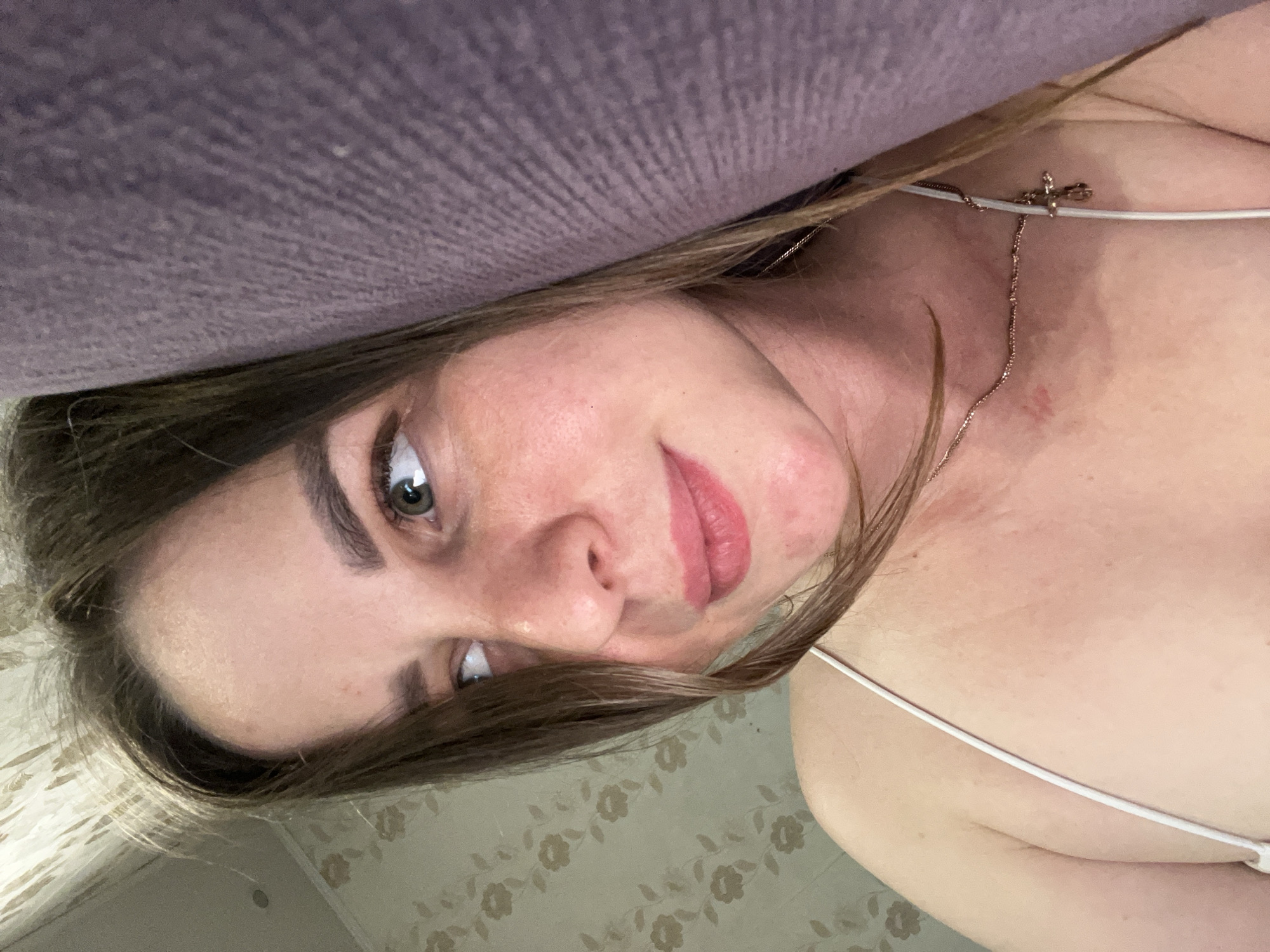 A close-up selfie captures a young woman indoors. She has long brown hair, which falls around her face, and prominent green eyes that look directly at the camera. Her lips are full and painted with a soft pink lipstick, and she has well-defined eyebrows. She wears a white spaghetti strap top, and a delicate gold-toned necklace with a small cross pendant is visible around her neck.
The image is tilted, creating a Dutch angle composition. A textured purple fabric, possibly a pillow or furniture, occupies the upper part of the frame, partially obscuring her hair. In the background, to the lower right, patterned wallpaper is visible. The wallpaper features a light-colored base with repeating, subtle floral or leafy motifs in a darker, muted tone. The lighting appears soft and even, suggesting an indoor setting without harsh sunlight. Her expression is calm and direct, possibly conveying engagement or mild curiosity. No discernible text is visible within the image. The setting appears to be a residential room in Izhevsk, Russia.