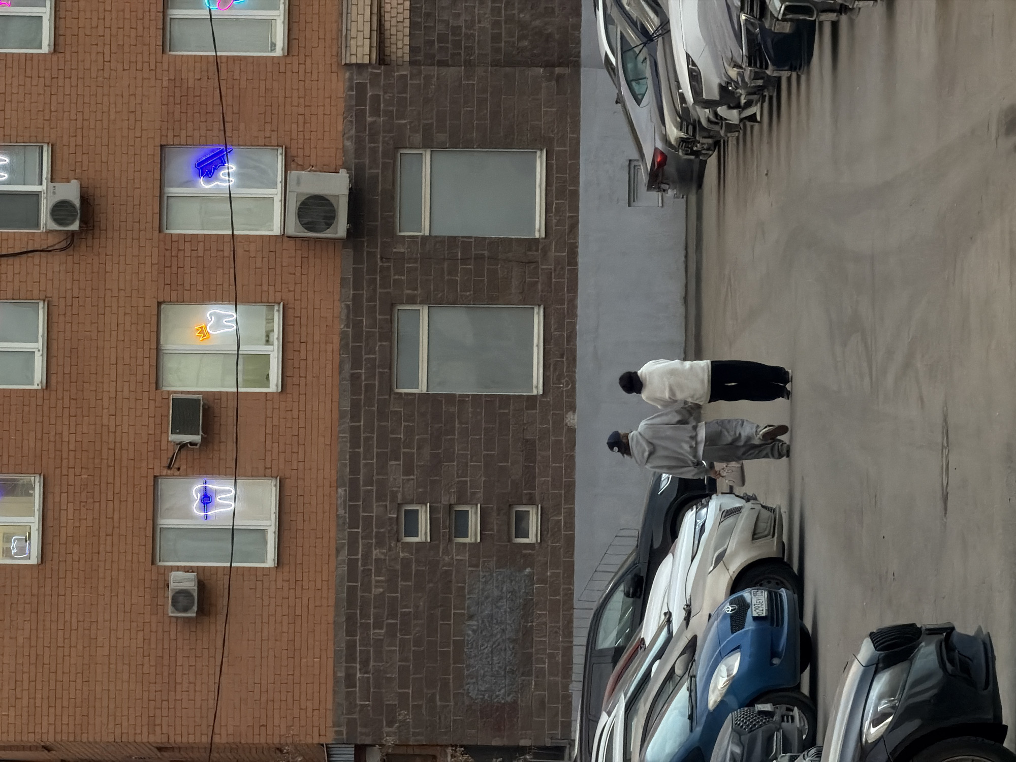 The image captures an urban scene in Khimki, Russia, featuring a multi-story brick building and a parking lot. The building's lower section is clad in dark, rough-textured brick, while the upper floors are constructed from red brick. Numerous rectangular windows with white frames are visible, and several air conditioning units are mounted on the red brick facade. Distinctive neon signs shaped like human teeth are illuminated in multiple upper-floor windows; one sign depicts a tooth with a gold crown, another features a tooth with blue braces, and a third shows a tooth with a blue dental tool or toothbrush.
In the foreground and midground, a paved parking area extends into the distance, flanked by rows of parked vehicles. Cars, predominantly sedans and SUVs of various makes and colors, are aligned on both the left and right sides. A blue Toyota Yaris, displaying a Russian license plate "O293EY 199," is visible among the vehicles on the left.
Two individuals are walking away from the camera down the central lane of the parking area. The person on the left is wearing a grey hooded sweatshirt, grey pants, and a dark baseball cap, carrying a light-colored handbag in their left hand. The person on the right is dressed in a light-colored, possibly off-white, long-sleeved top or jacket and dark pants. They are walking side-by-side, moving towards the background of the scene.