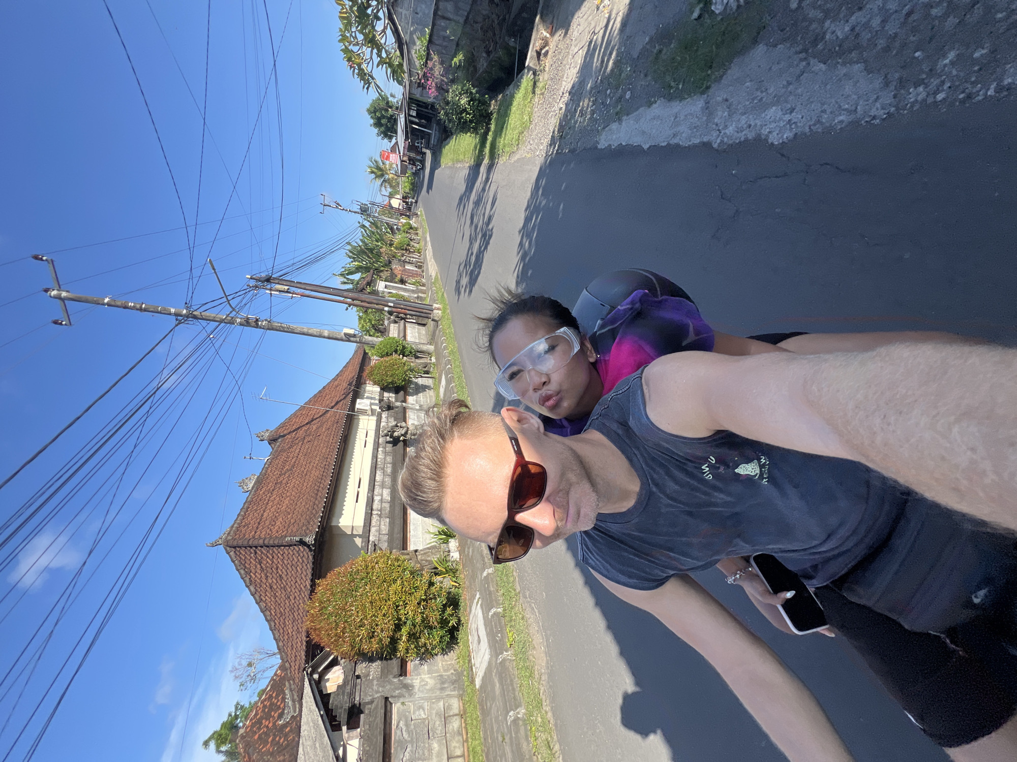 The photograph depicts a man and a woman riding on a scooter down a road. The man, wearing sunglasses and a dark tank top, is driving the scooter and holding a phone in his left hand. The woman, wearing safety goggles and a helmet, is riding behind him. They appear to be taking a selfie. The background features a road, buildings with traditional Balinese architecture, and utility poles with wires. The sky is clear and blue. Based on the architectural style and other contextual clues, the location is likely Sukawati, Indonesia.